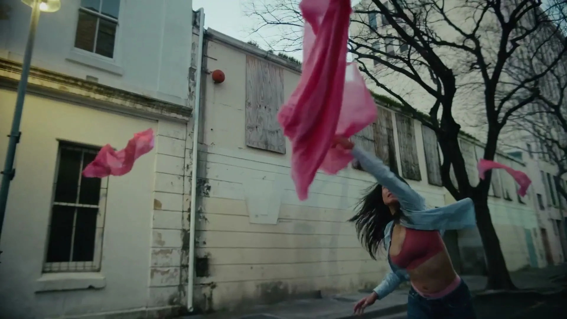 A young woman in a bra and jeans runs down a street, reaching for a flowing pink fabric that billows around her.