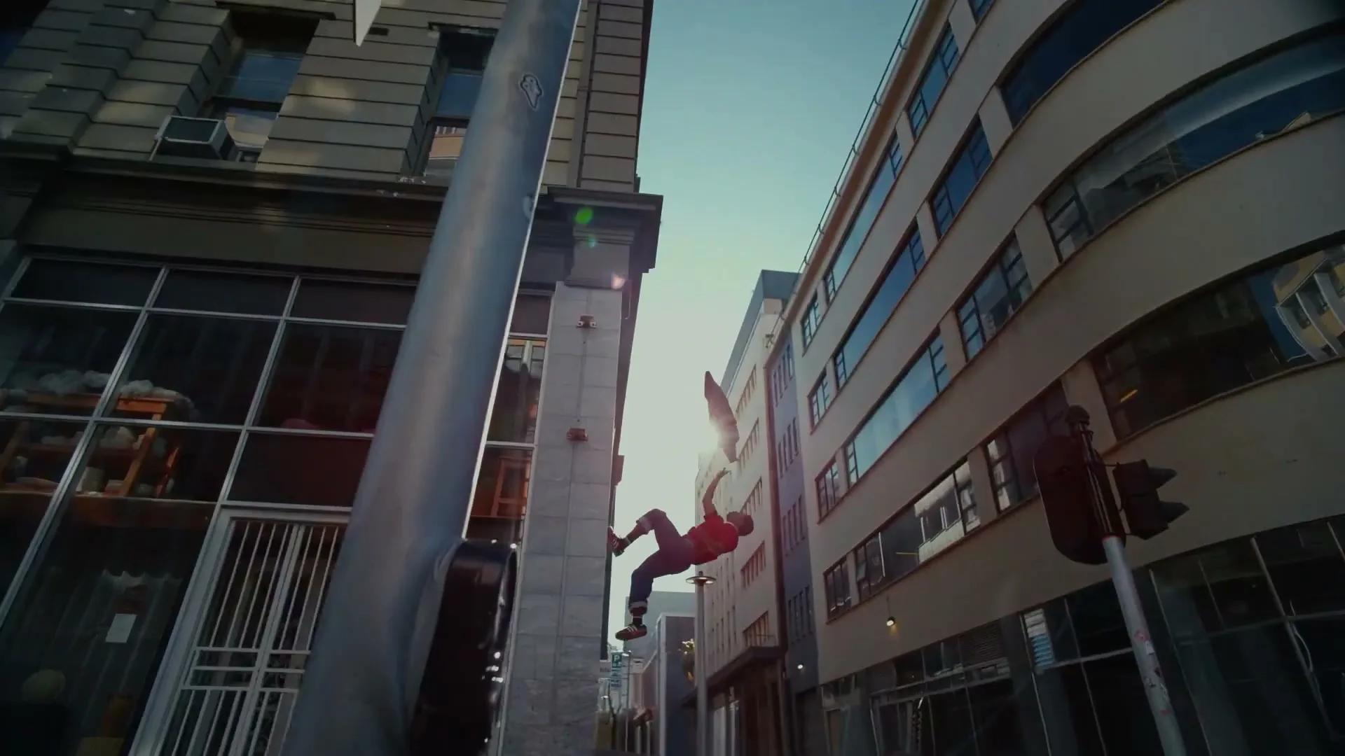 A male in a red shirt and blue pants is seen falling backwards through the air between two buildings on a city street during the day.