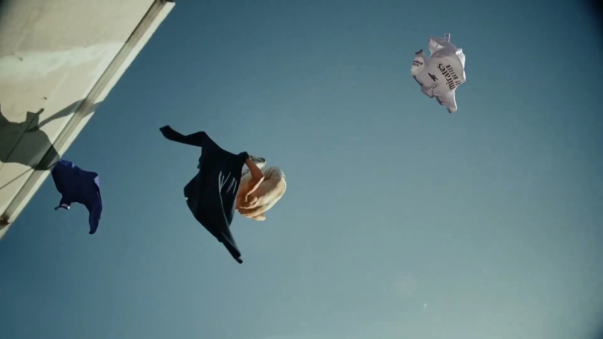 A male dancer in his 20s performs a backflip in mid-air, surrounded by floating clothes against a clear blue sky.