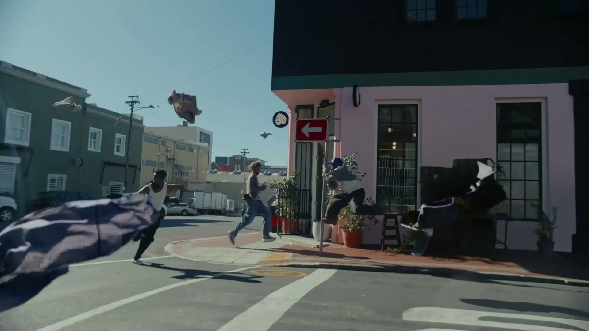 Three young men run through a city street as clothes fly around them, with a pink building on the right.