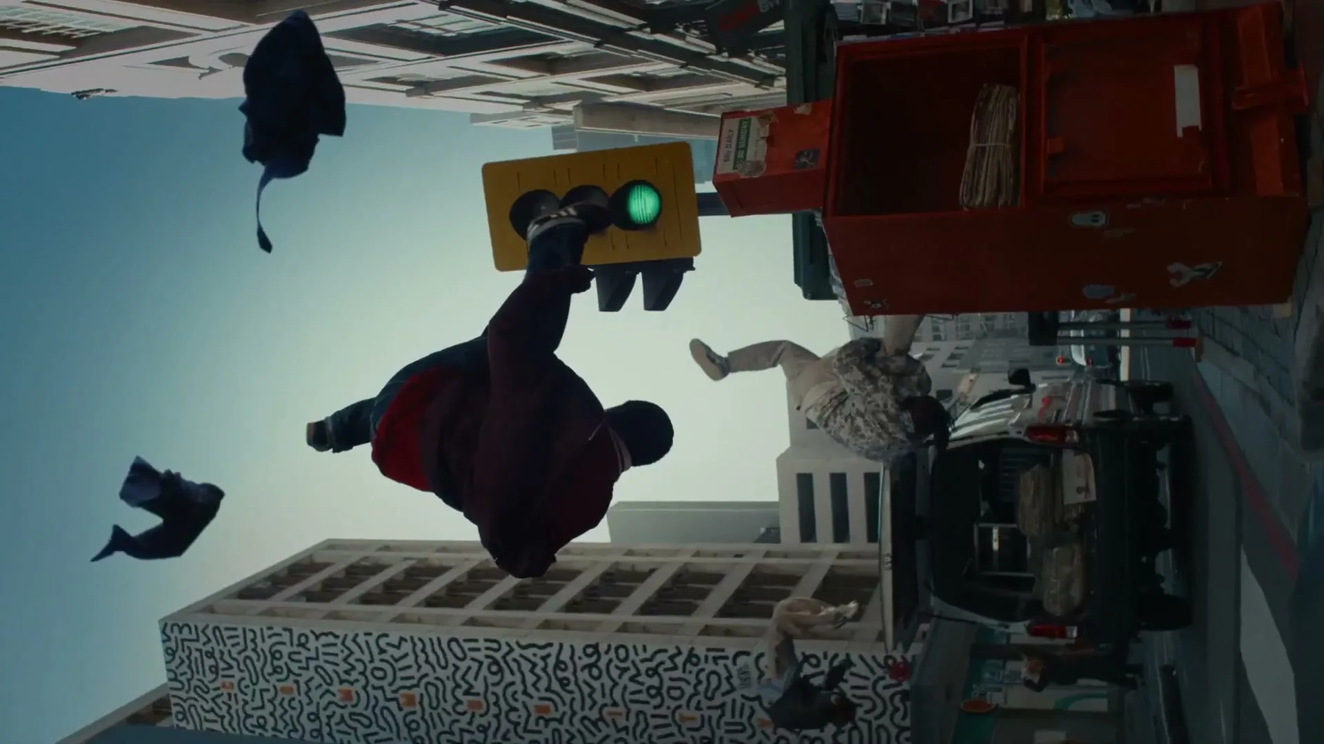 A man in a red jacket and black pants floats upside down in a city street, with a traffic light and buildings also inverted, as if gravity has reversed.