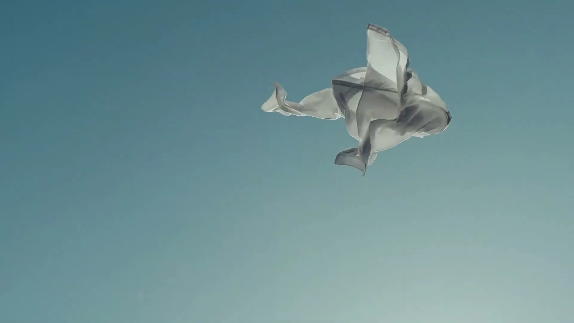 A light grey fabric piece floats gracefully against a clear blue sky, gently swaying in the air.