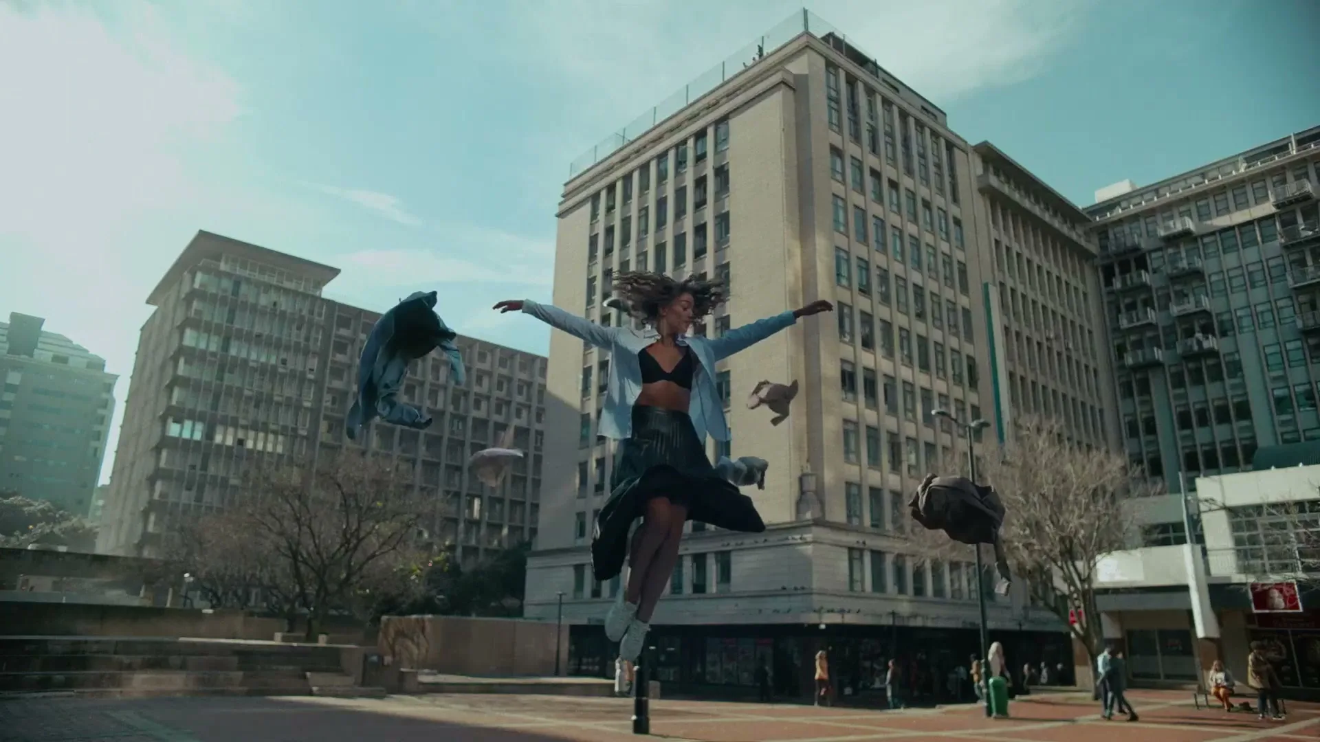 A young woman with curly hair floats in the air with arms outstretched, surrounded by floating clothes and a backpack, in an urban plaza with tall buildings.