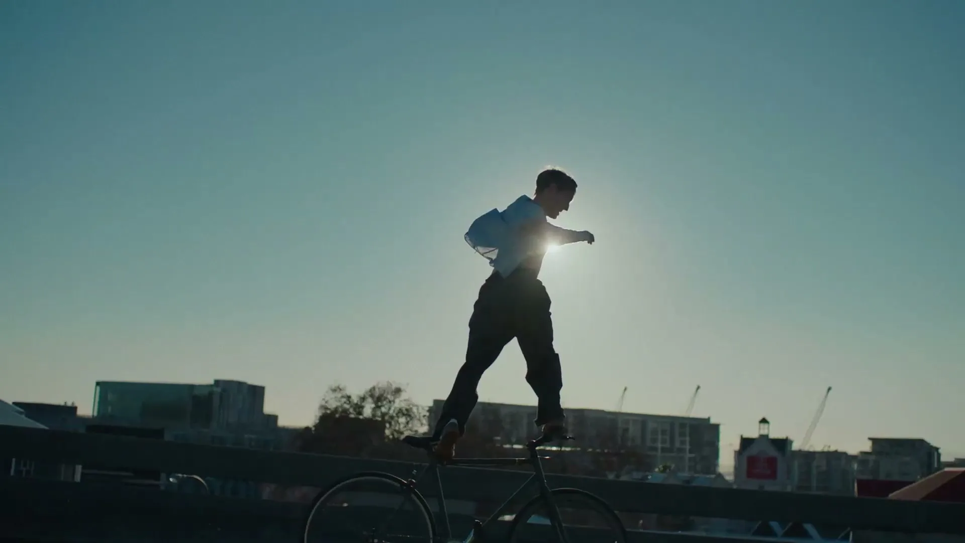 A person in silhouette stands on a bicycle, balancing against a bright sun, with city buildings in the background.