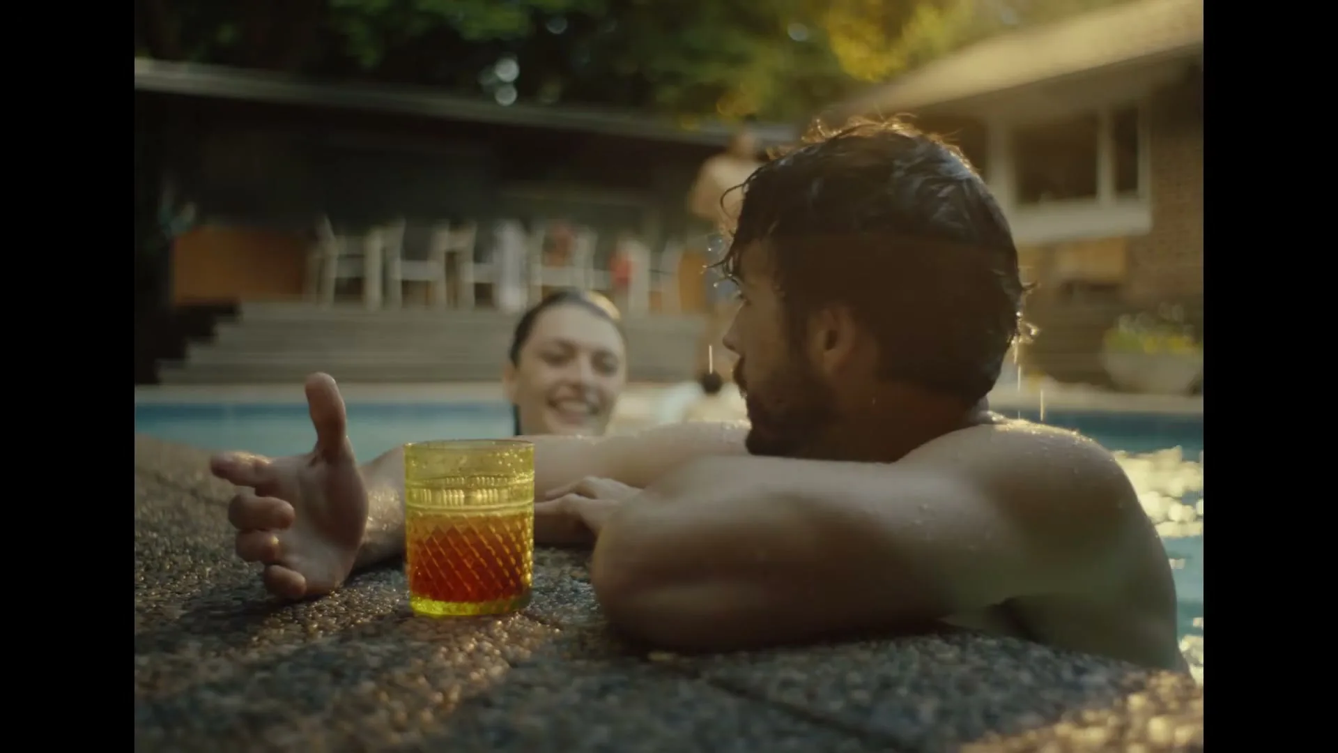 A man with dark hair and a beard is in a swimming pool, holding onto the edge, with a drink next to him, as a woman splashes him playfully.
