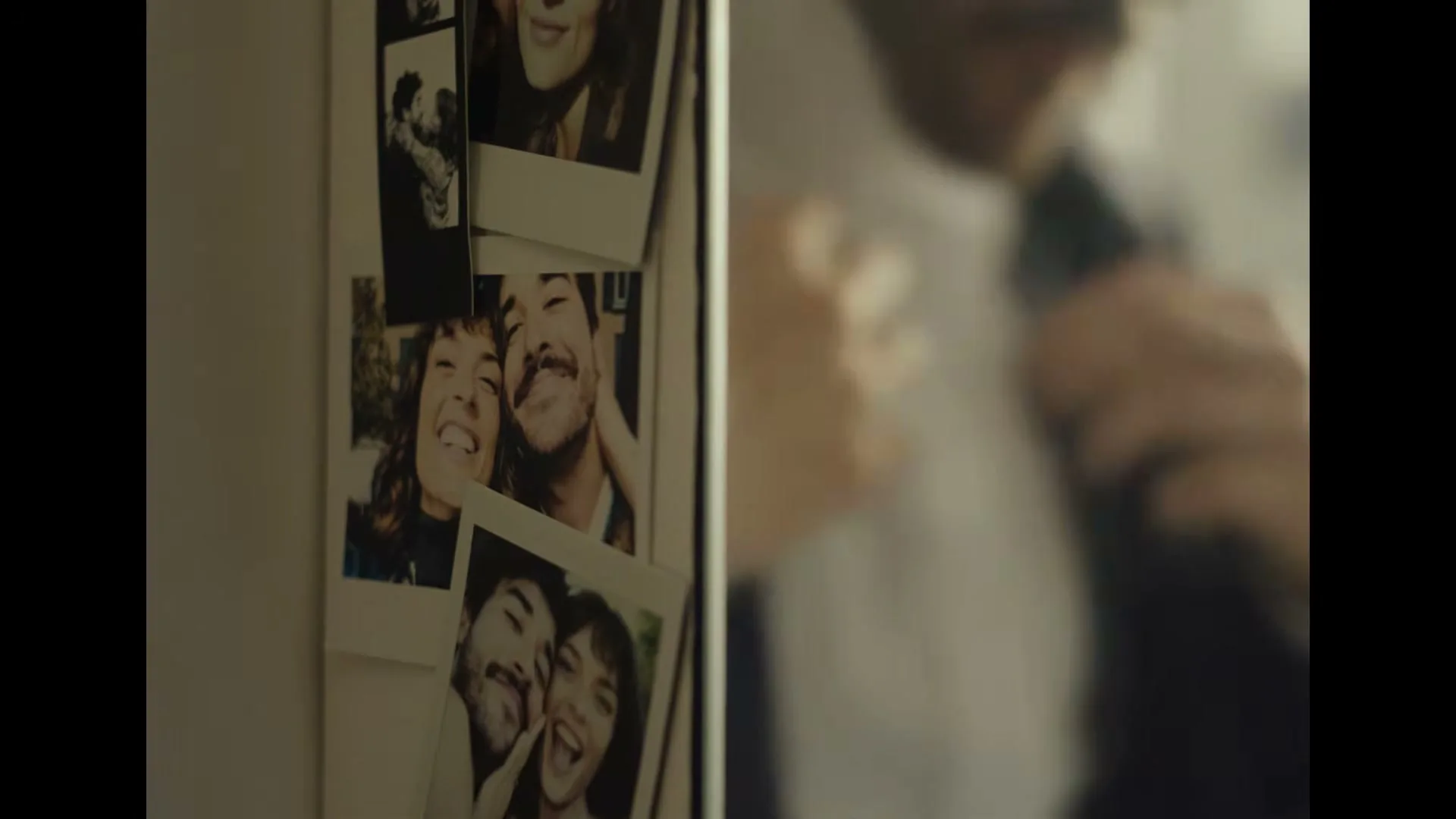 A man in a white shirt and dark tie is seen in soft focus, adjusting his tie, with several polaroid photos of a couple on a fridge in the foreground.