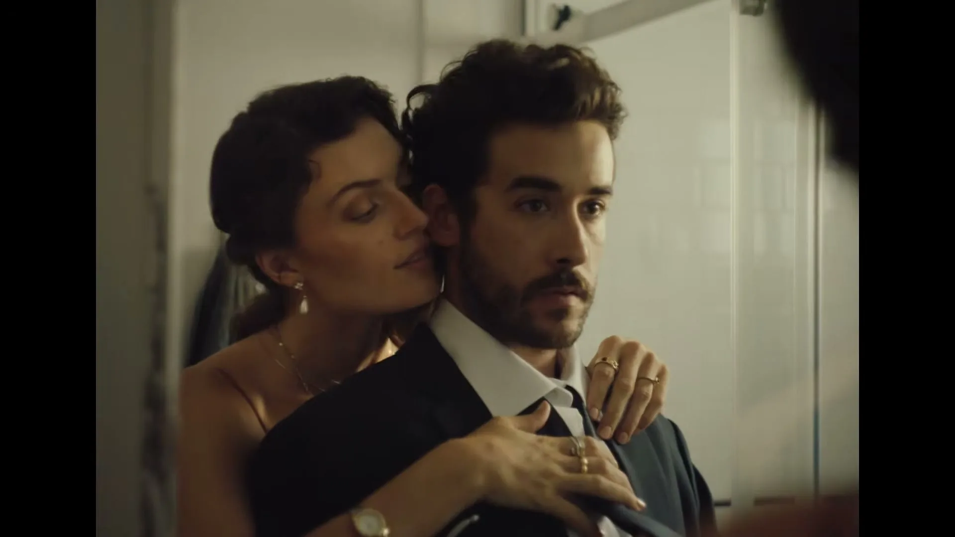 A woman in a formal dress embraces a man in a suit from behind as he adjusts his tie in a bathroom mirror.