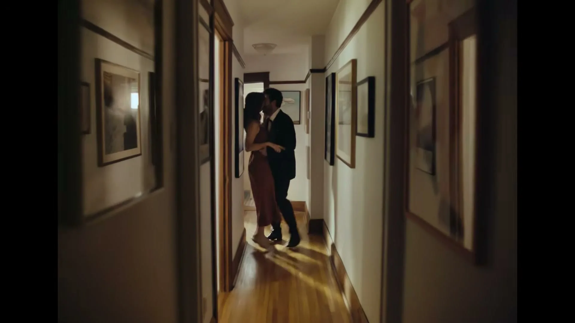 A couple, a man in a suit and a woman in a dress, embrace and kiss in a dimly lit hallway of a home.