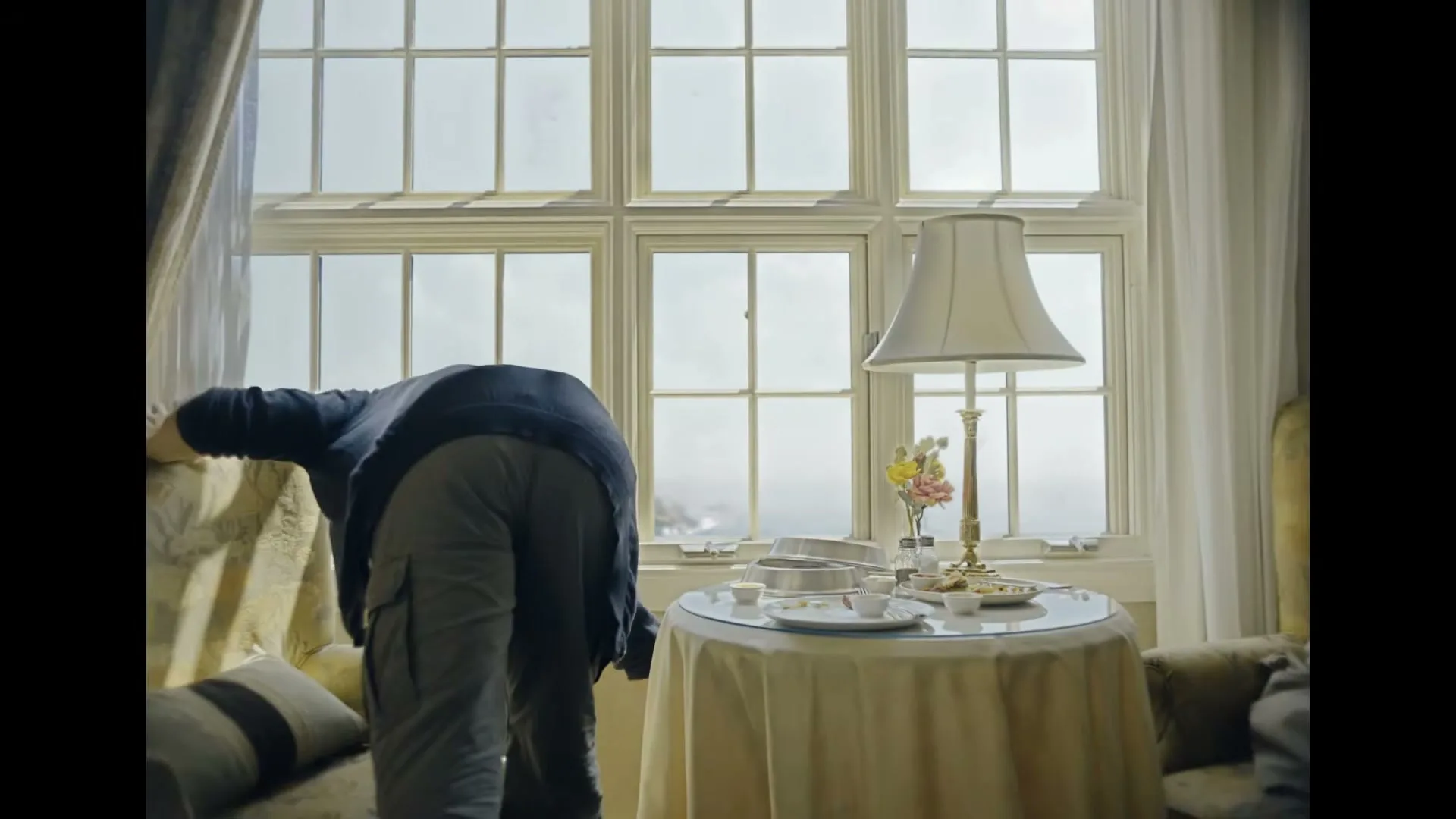 A man and woman frantically pack their belongings in a hotel room with a view of a lighthouse and the ocean.