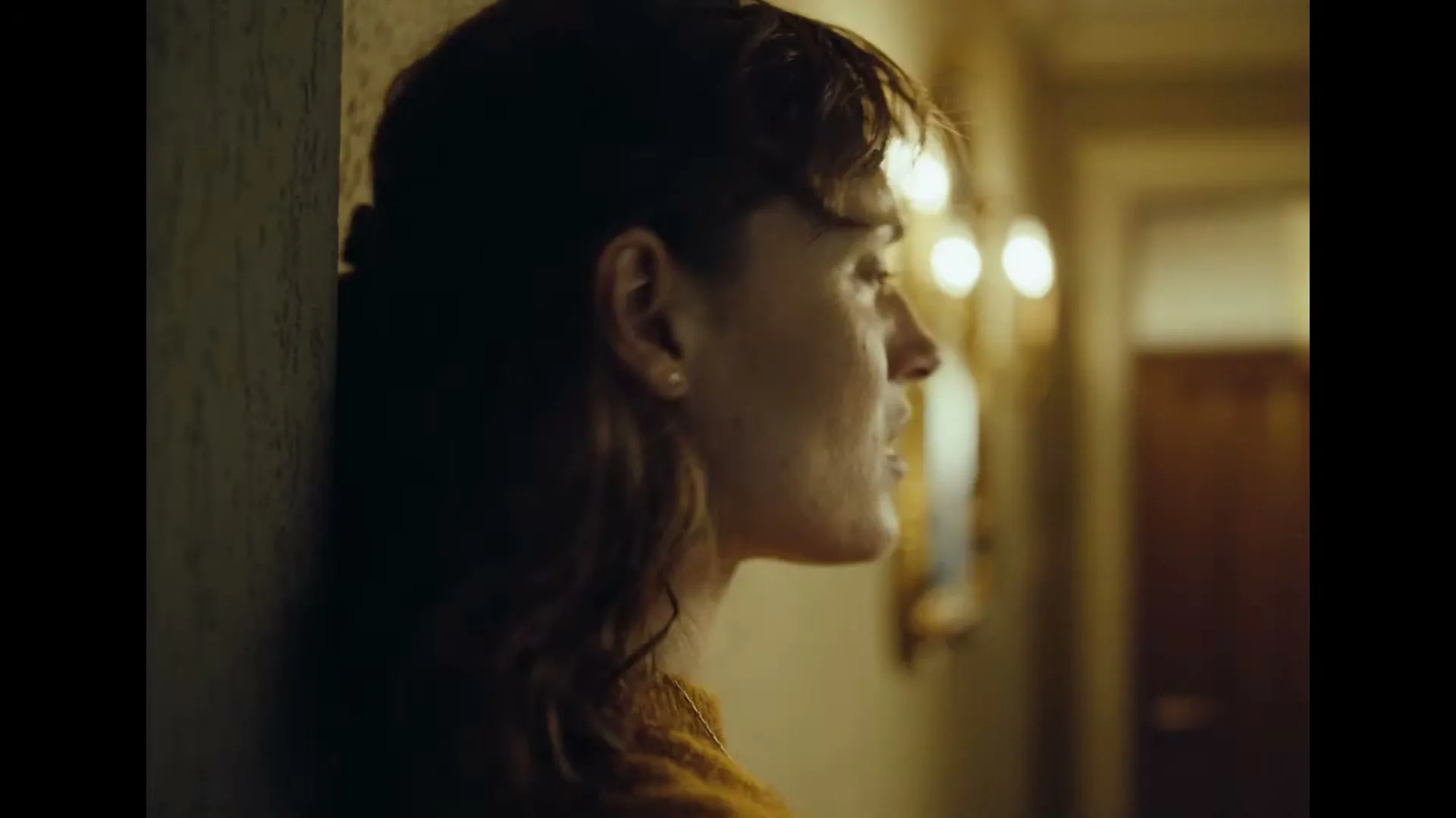 A young woman with brown hair and a mustard yellow sweater leans against a textured wall in a dimly lit hallway, looking concerned.