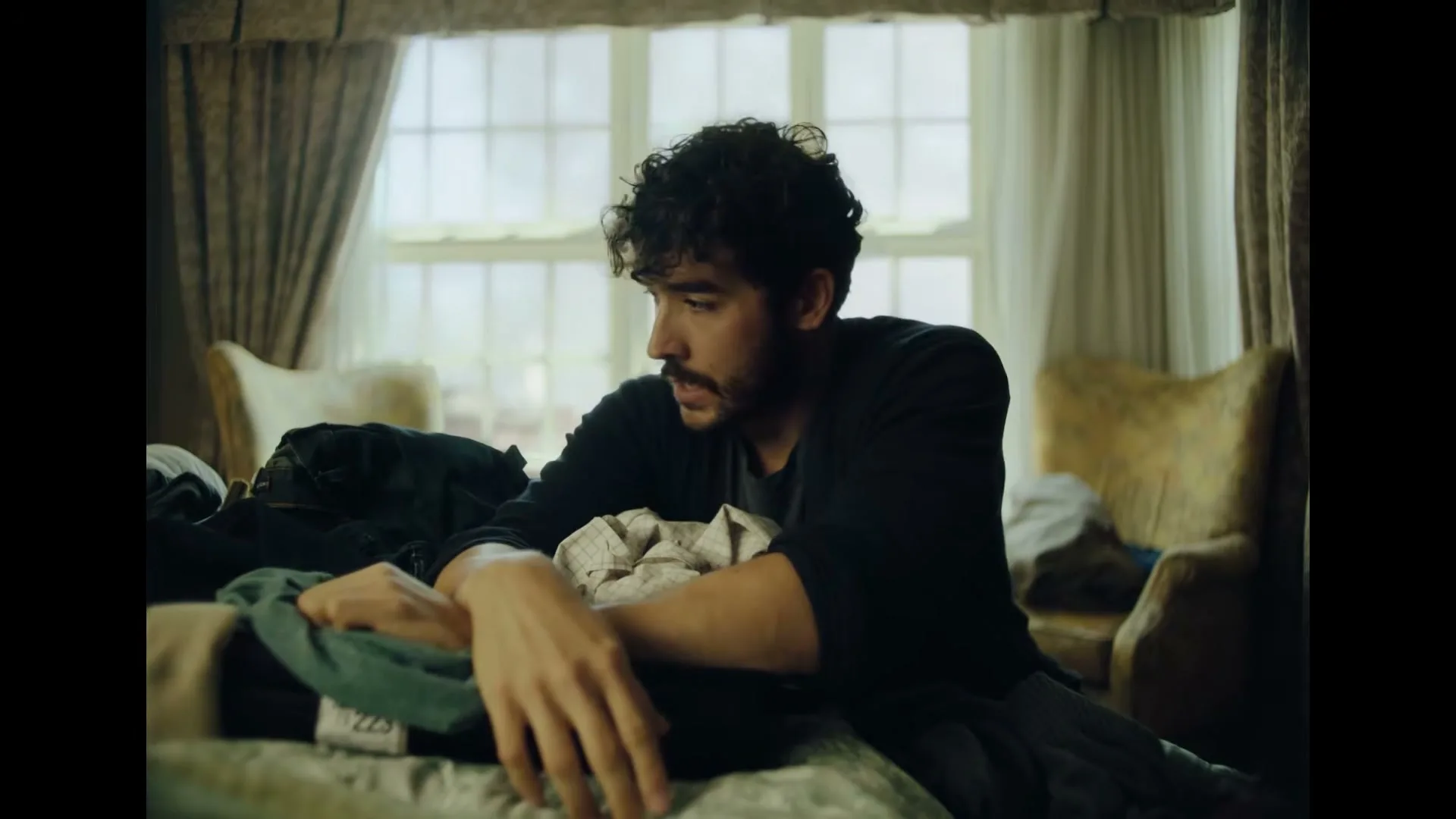 A man with dark curly hair and a beard sits on a bed, looking down at a pile of clothes in a bedroom with a large window.