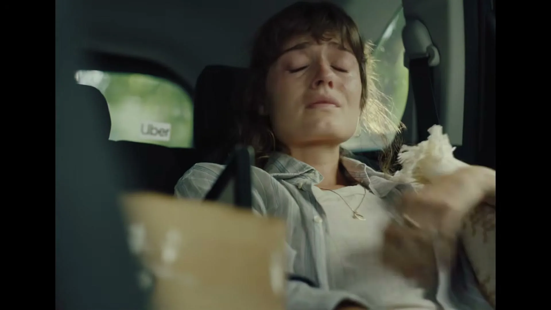 A young woman with brown hair, wearing a light shirt, sits in the back of a car, looking distressed and covering her mouth with her hand.
