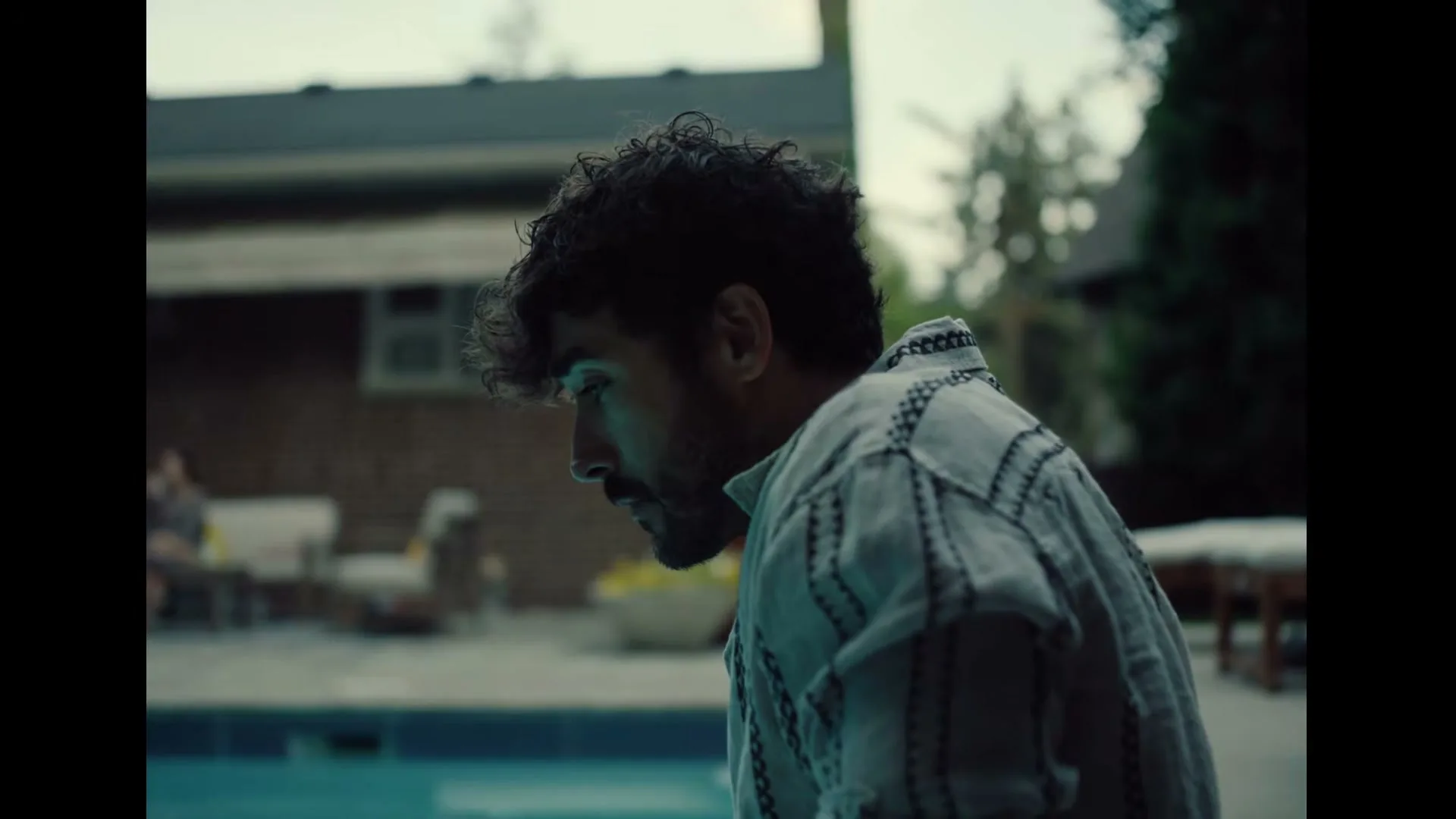 A man with curly hair and a patterned shirt looks down at a swimming pool in a backyard during the day.