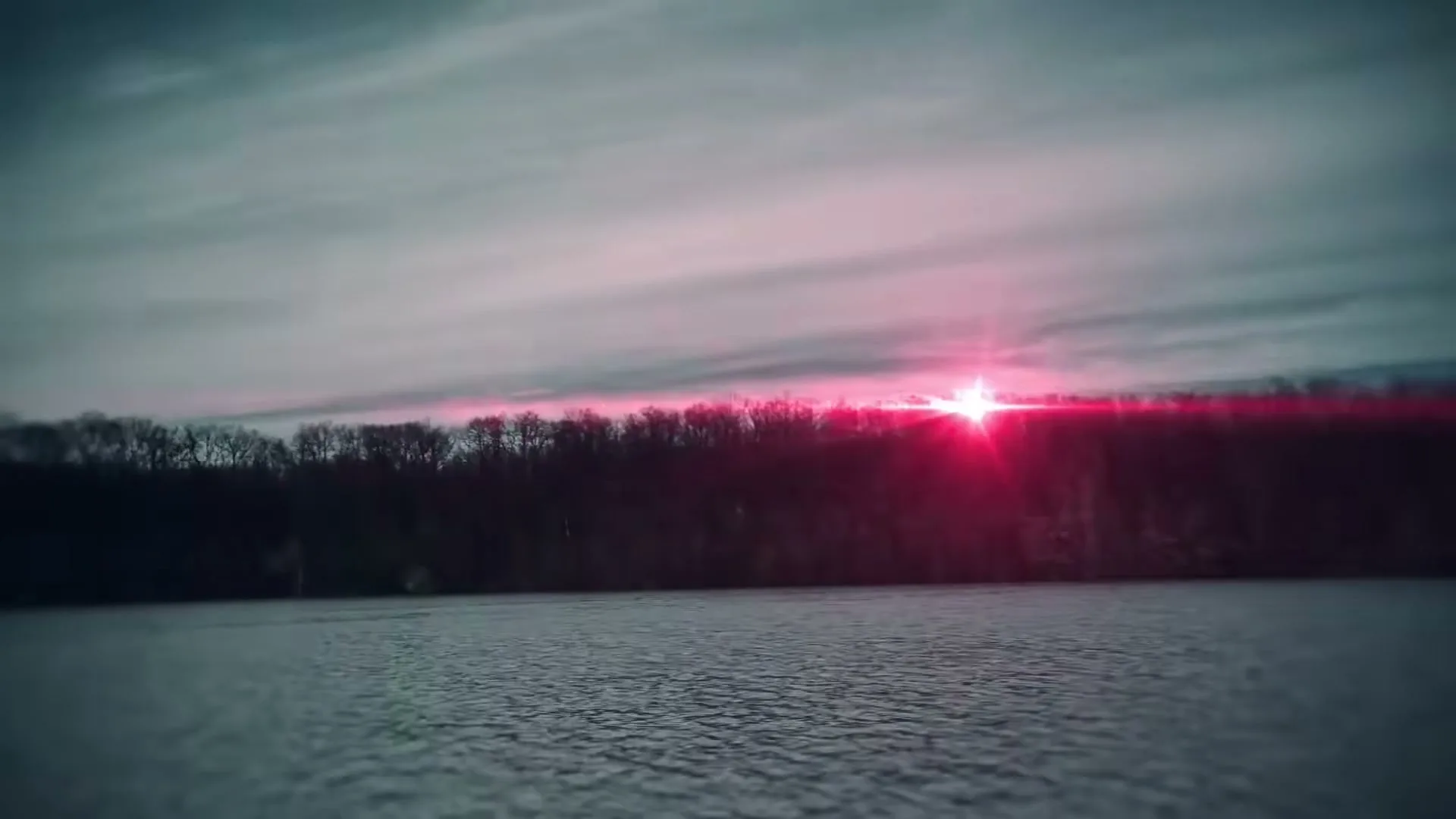 A wide shot of a lake with a dark tree line under a cloudy sky, featuring a bright pink lens flare from the setting sun.