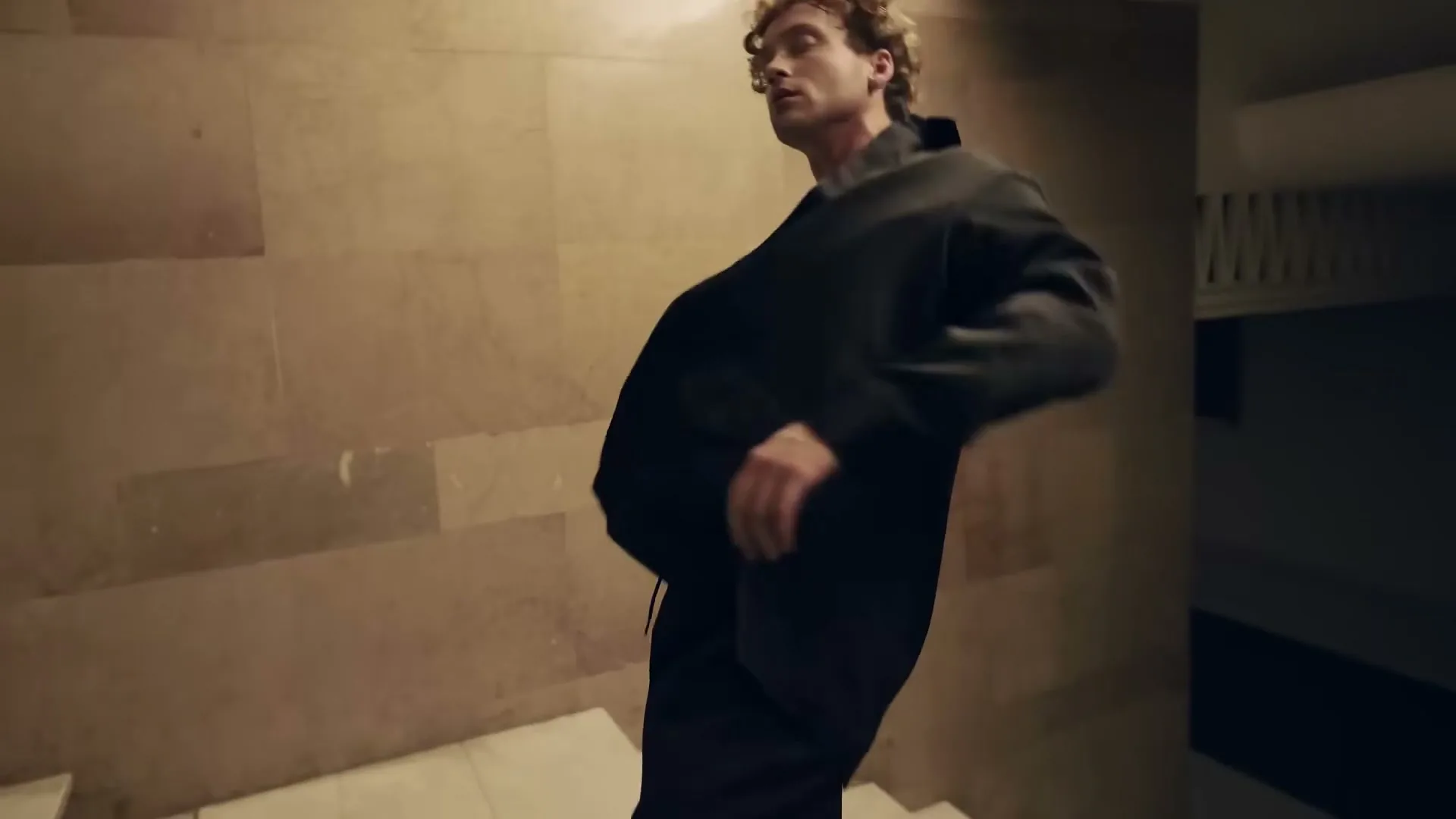 A young man with curly hair walks up a staircase, turning to look at the camera as his jacket falls open, revealing a white shirt.