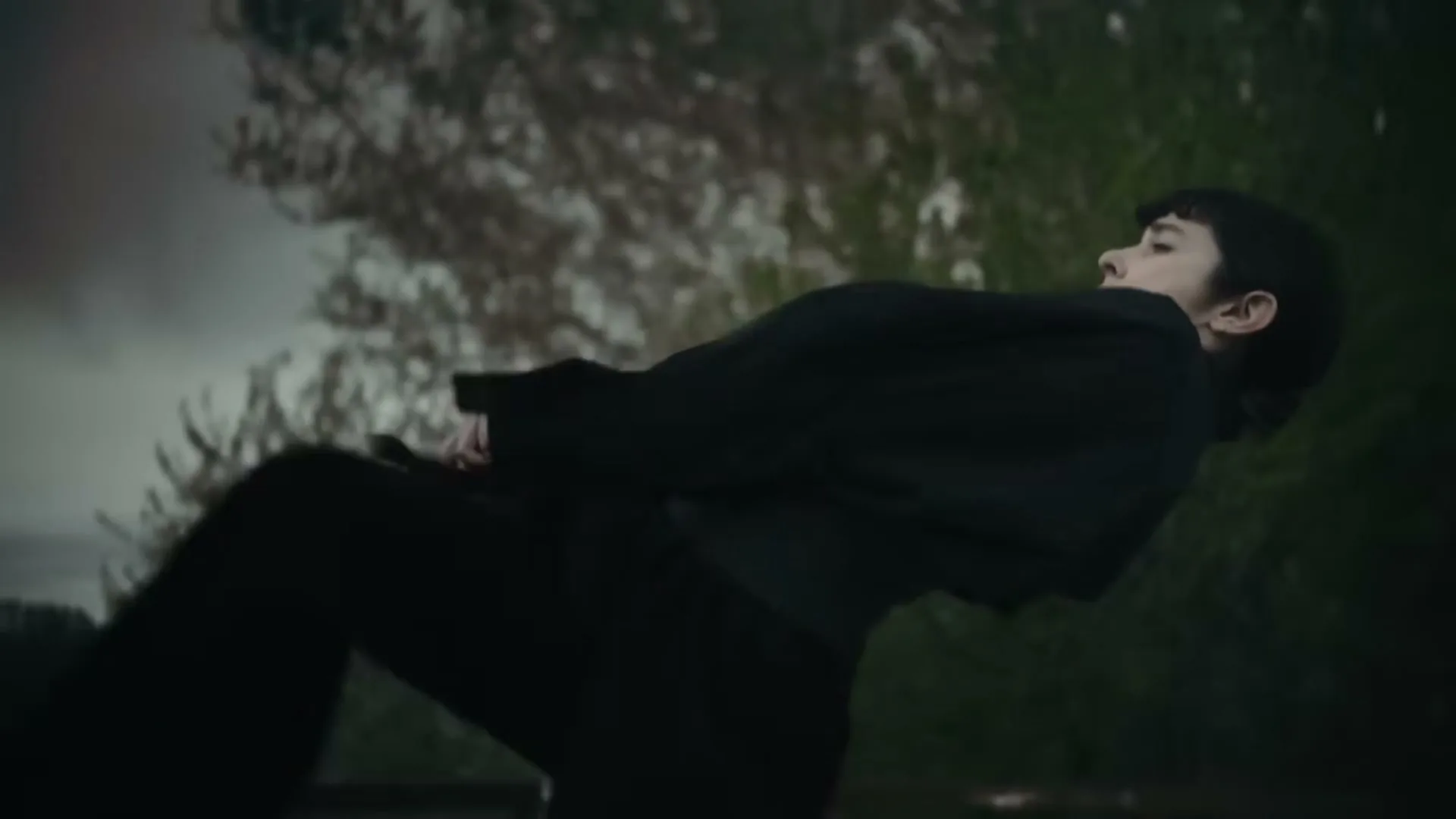 A young person in a black suit is dramatically falling backward with arms outstretched, against a blurred outdoor background with trees.