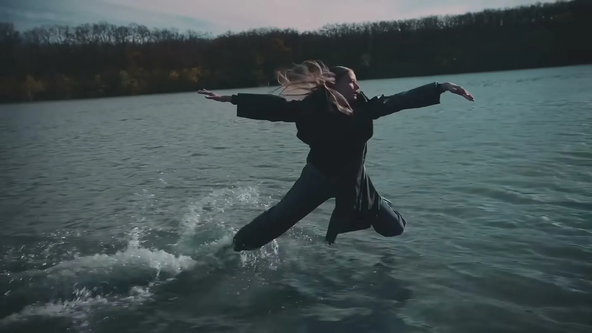 A young woman in dark clothing leaps gracefully over the surface of a lake, creating a splash, with trees in the background.