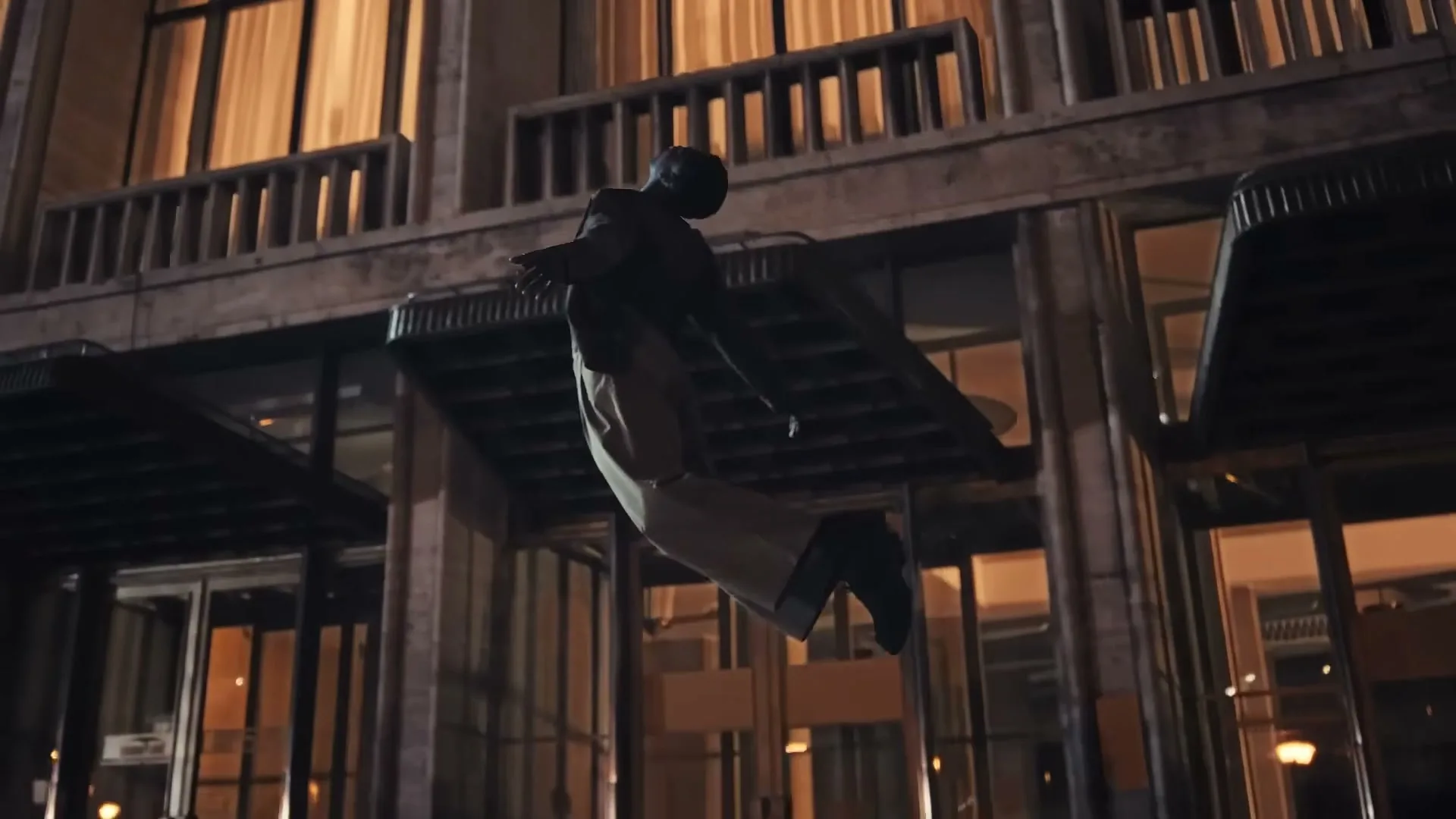 A person in a dark suit and light pants is suspended in mid-air, floating in front of a modern building with illuminated windows at night.