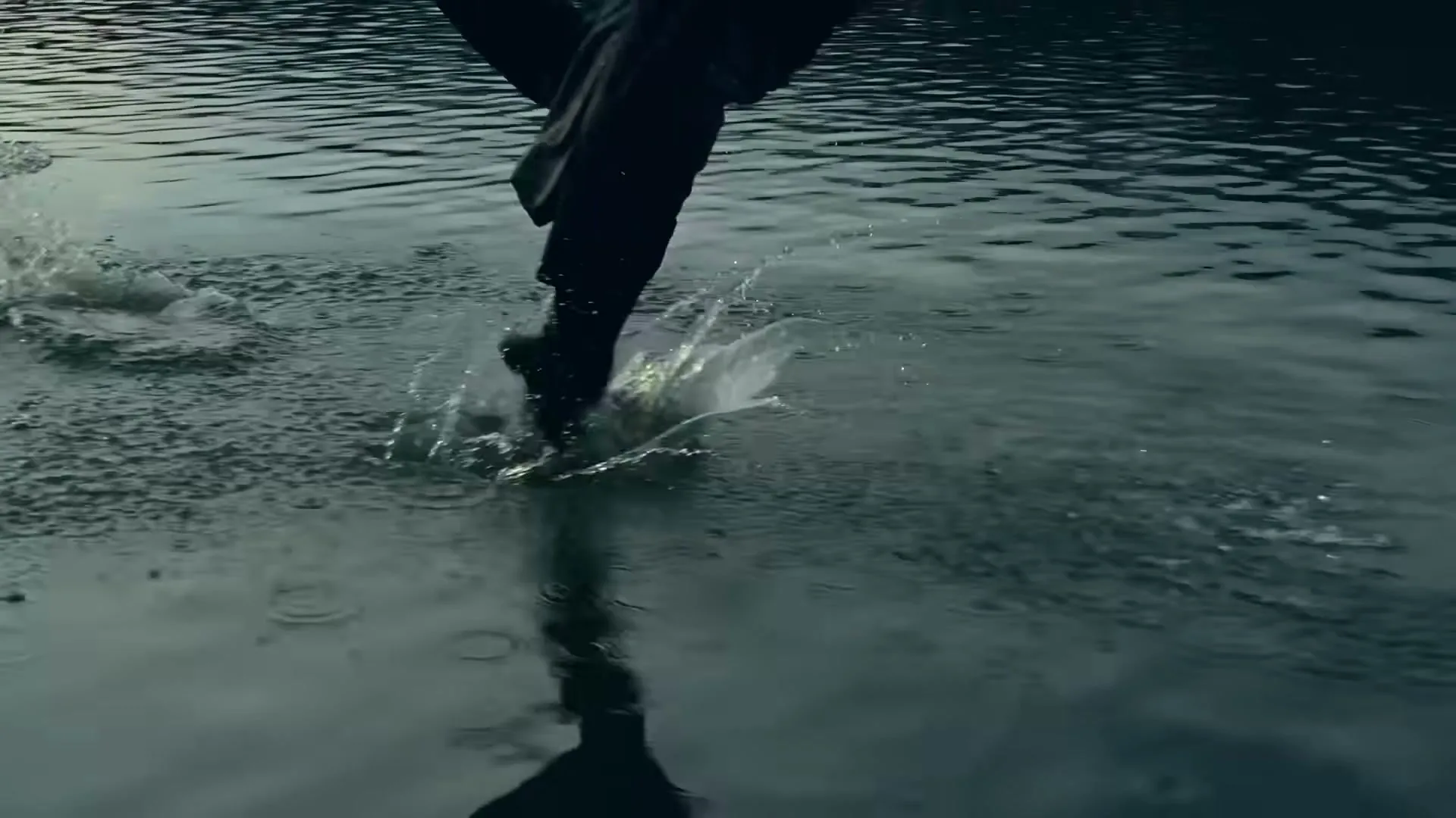 A person in dark clothing runs through shallow water, creating splashes, with their legs and feet visible from the knees down.