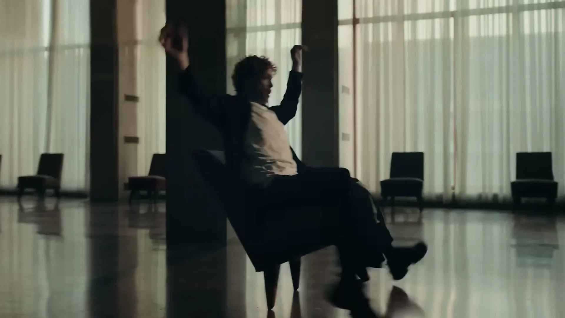 A man in a dark suit and white shirt falls backward into a chair in a large, empty room with tall windows.