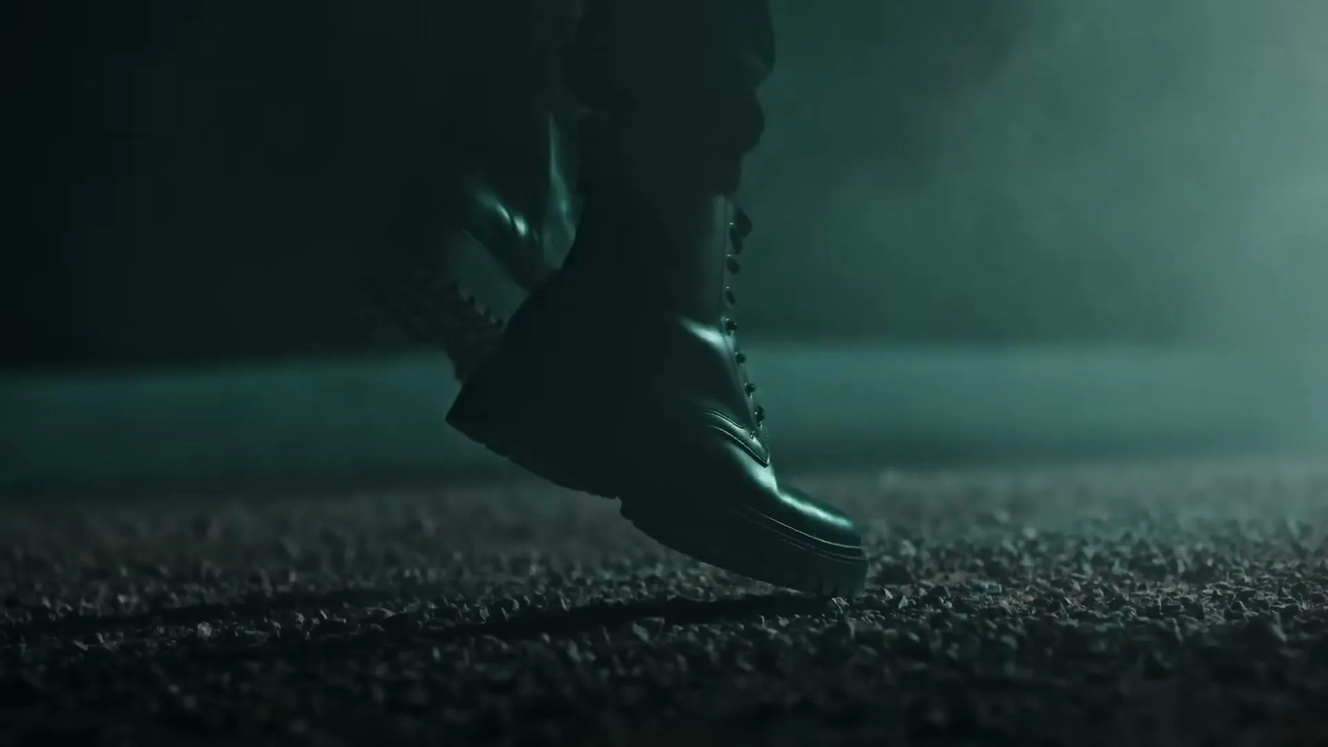 A person wearing black boots walks on a gravelly surface in a dark, smoky environment with a green light source.
