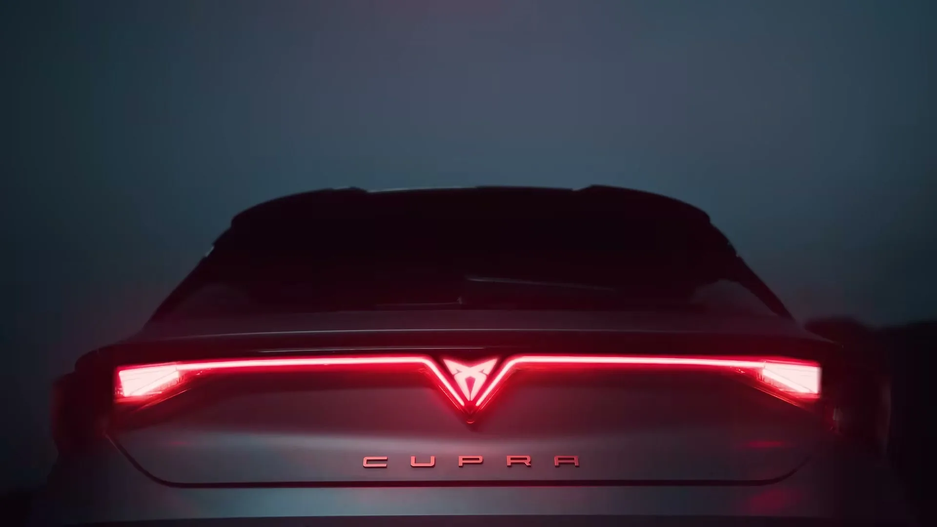 The rear of a dark grey Cupra car is shown in a medium shot, with its red tail lights illuminating in a dark, indeterminate setting.