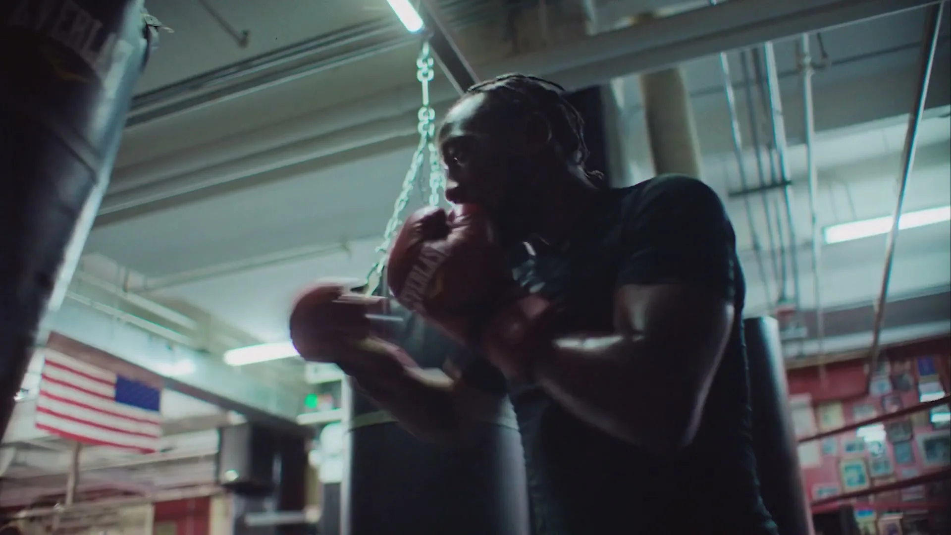 A male boxer with dreadlocks and red gloves trains with a punching bag in a dimly lit gym, hitting the bag repeatedly.