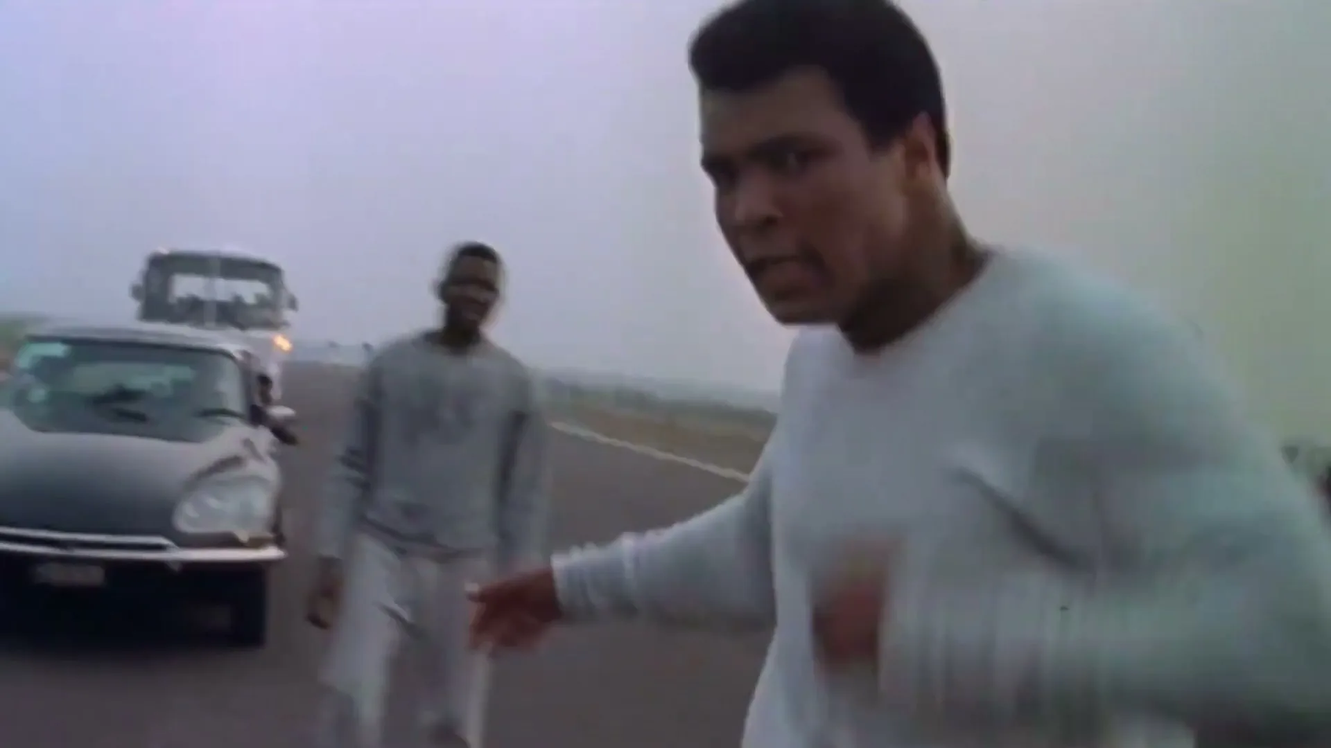 Muhammad Ali, wearing a white sweatshirt, shadowboxes on a misty road with other men and vehicles in the background.