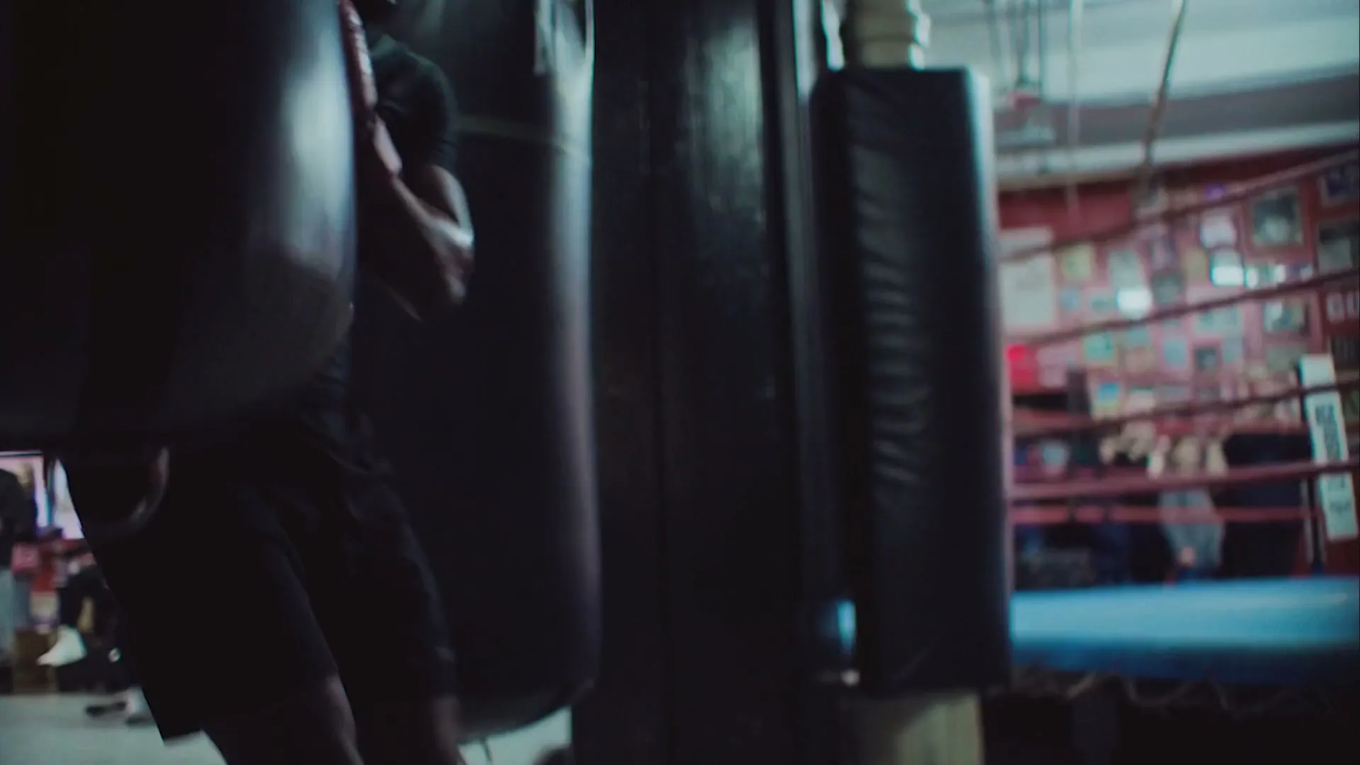 A male boxer in red gloves trains with a punching bag in a dimly lit boxing gym, with other people visible in the background.