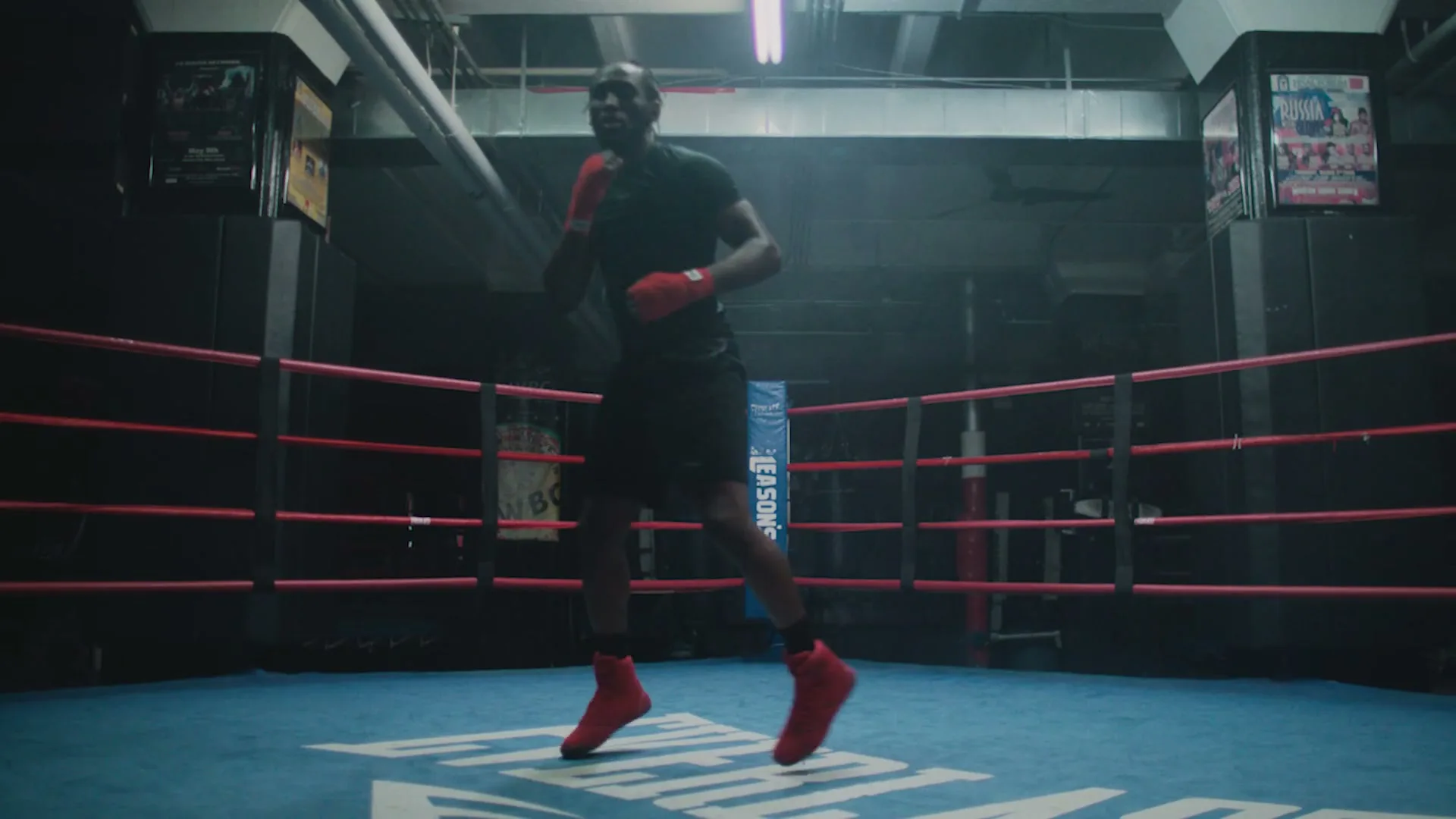 A male boxer in black attire and red gloves shadowboxes in a boxing ring in a dimly lit gym.