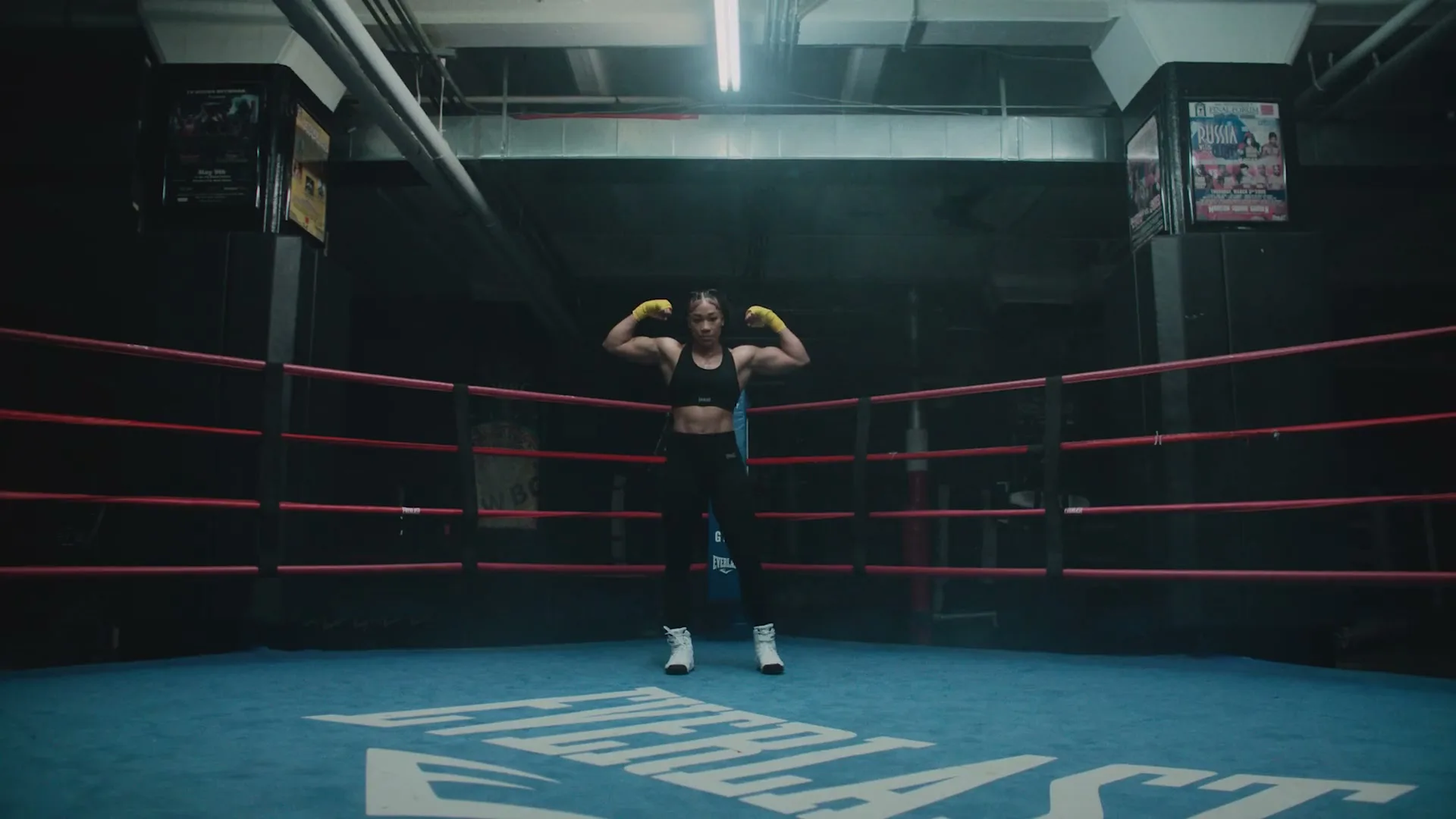 A female boxer in a black sports bra and leggings flexes her biceps in a boxing ring.