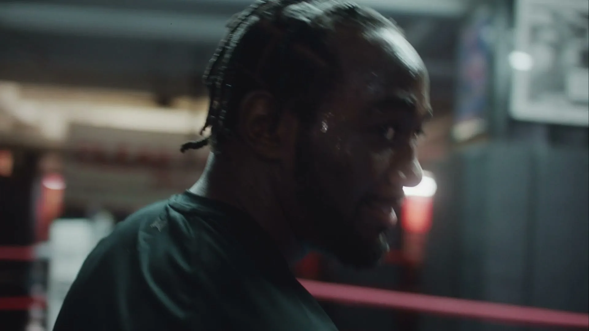 A sweaty Black man with braided hair, wearing a black t-shirt, turns his head and smiles in a boxing gym.