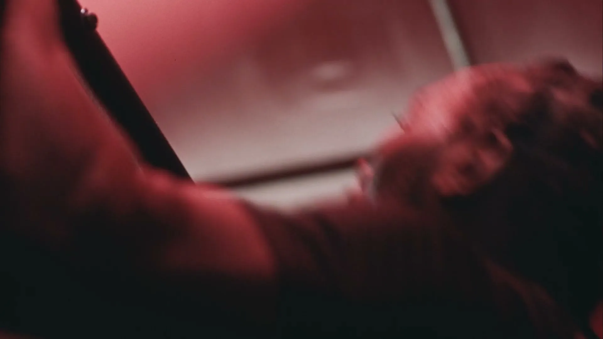 A man with braided hair, wearing a dark shirt, is seen from a low angle, looking up and moving his head, in a red-lit interior.