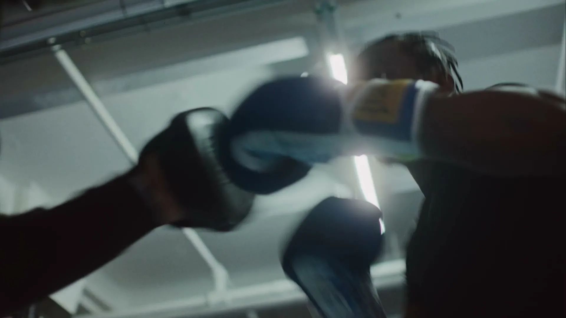 A male boxer with dreadlocks, wearing blue boxing gloves, practices punching a pad held by a trainer in a dimly lit gym.