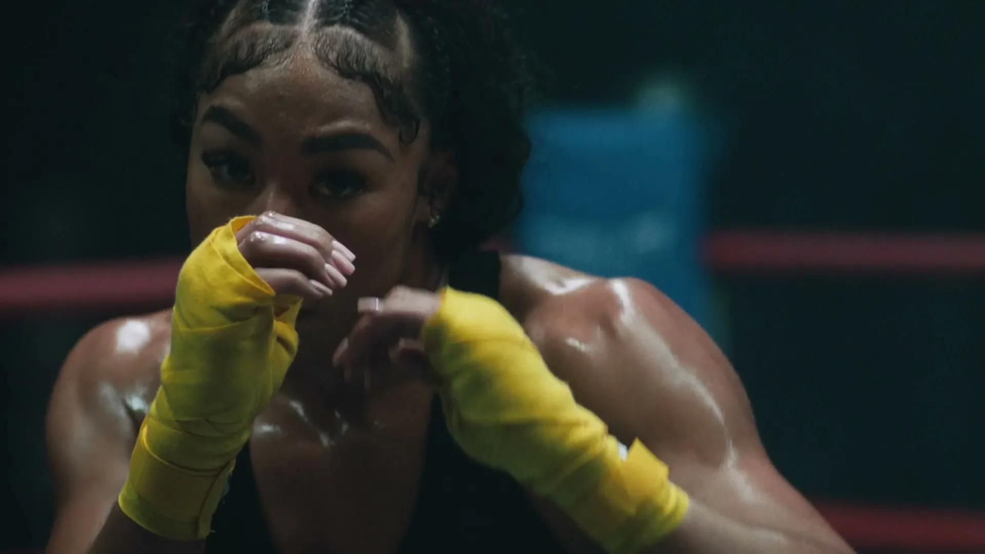 A determined female boxer with braided hair and yellow hand wraps trains in a boxing ring, throwing punches with sweat glistening on her skin.