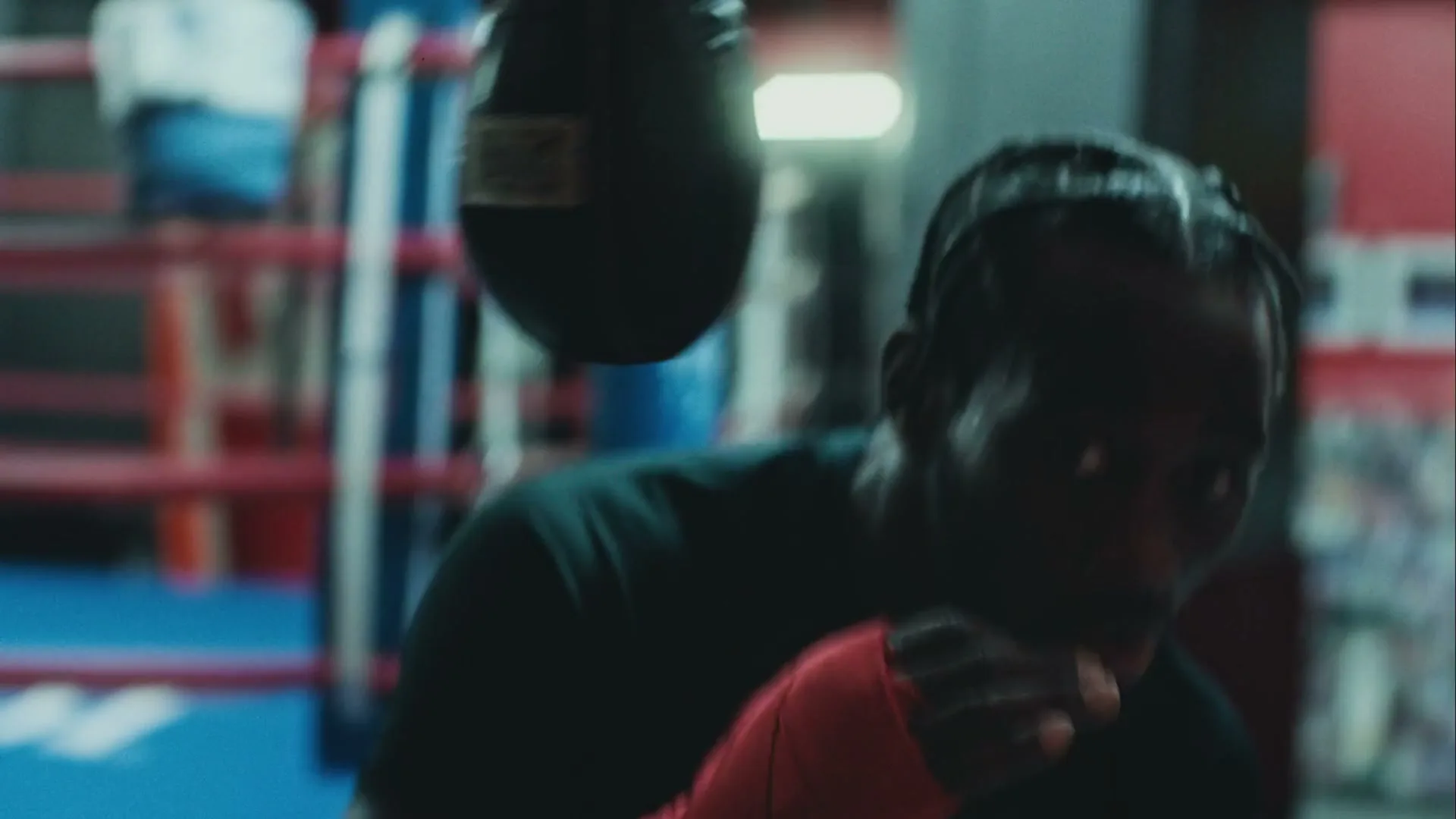 A male boxer with braided hair and red hand wraps trains with a speed bag in a boxing gym.