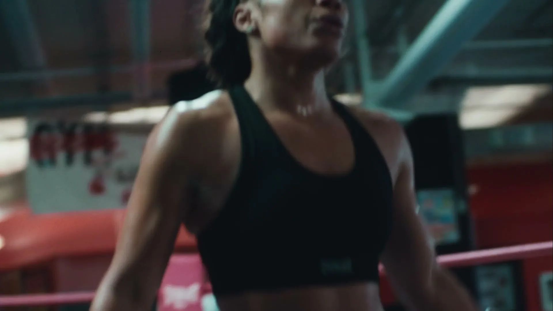 A sweaty female boxer in a black sports bra is jumping rope in a boxing ring at a gym, looking determined.