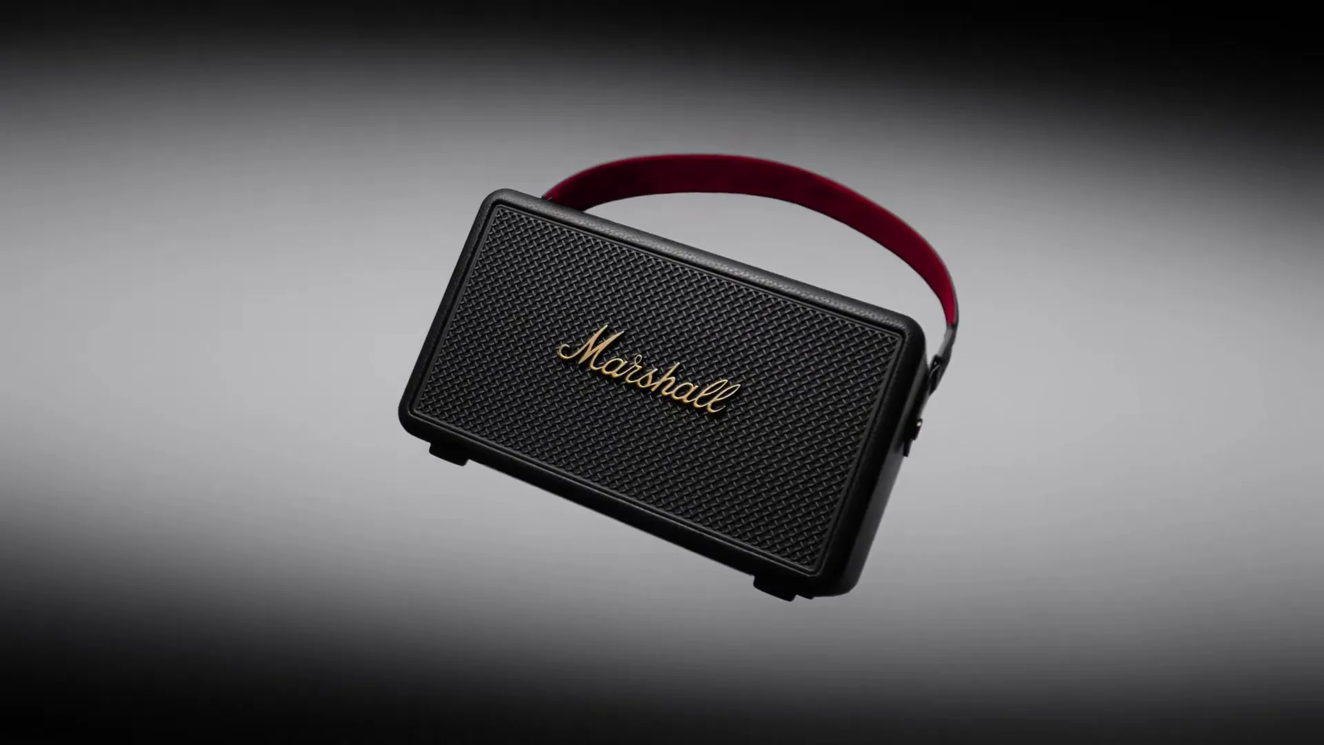 A black Marshall Kilburn III portable speaker with a red strap rotates against a dark grey background, showcasing its design.