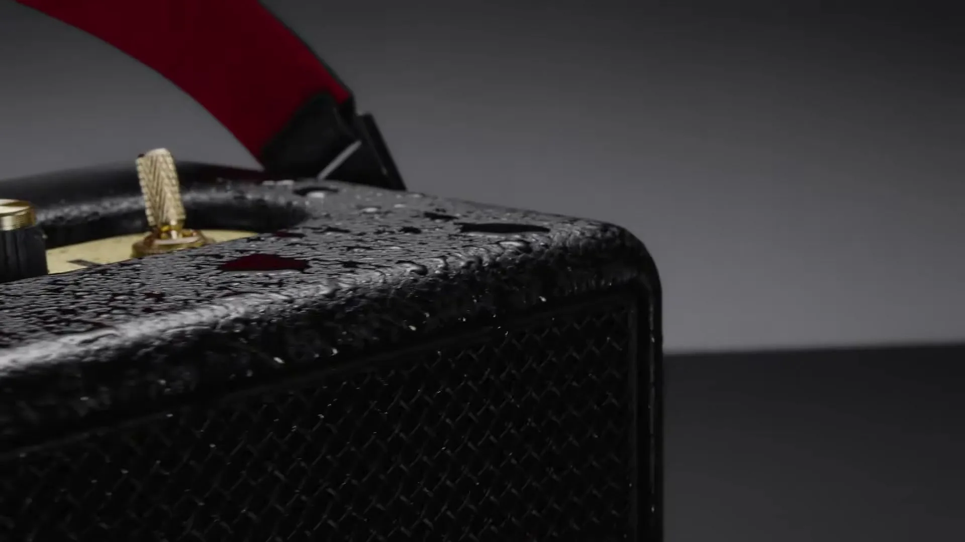 A black Marshall speaker with a red strap and gold accents is covered in water droplets, slowly rotating against a dark background.