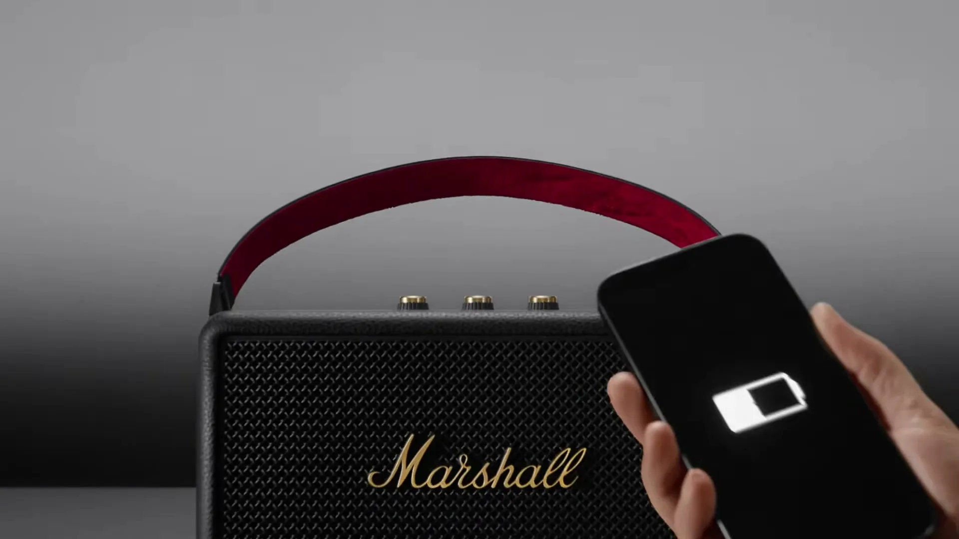 A person's tattooed arm holds a smartphone with a low battery icon, plugging it into a black Marshall speaker with a red strap on a grey surface.
