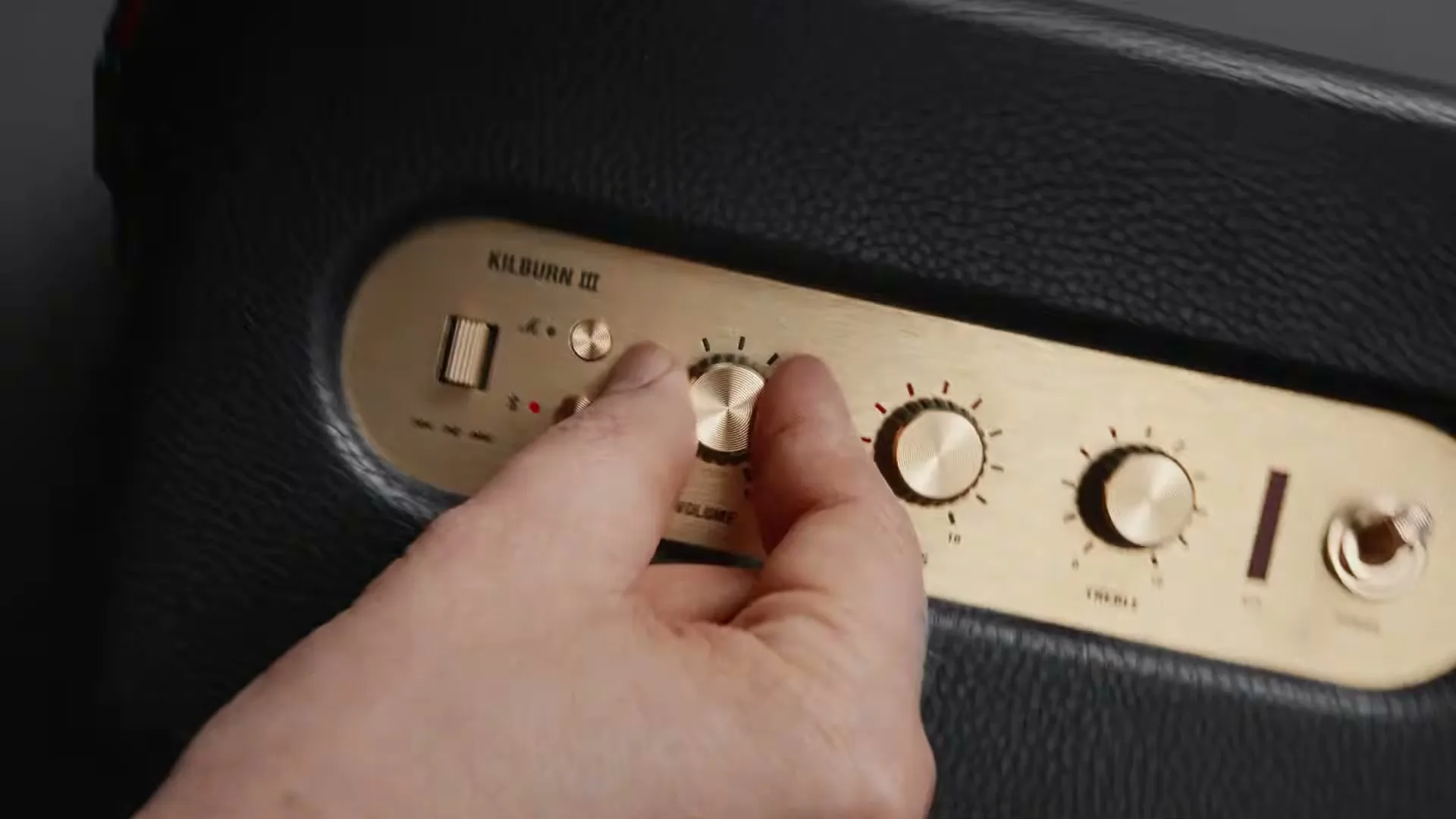 A person's hand adjusts the volume knob on a black and gold Marshall speaker, revealing more of the speaker's control panel.