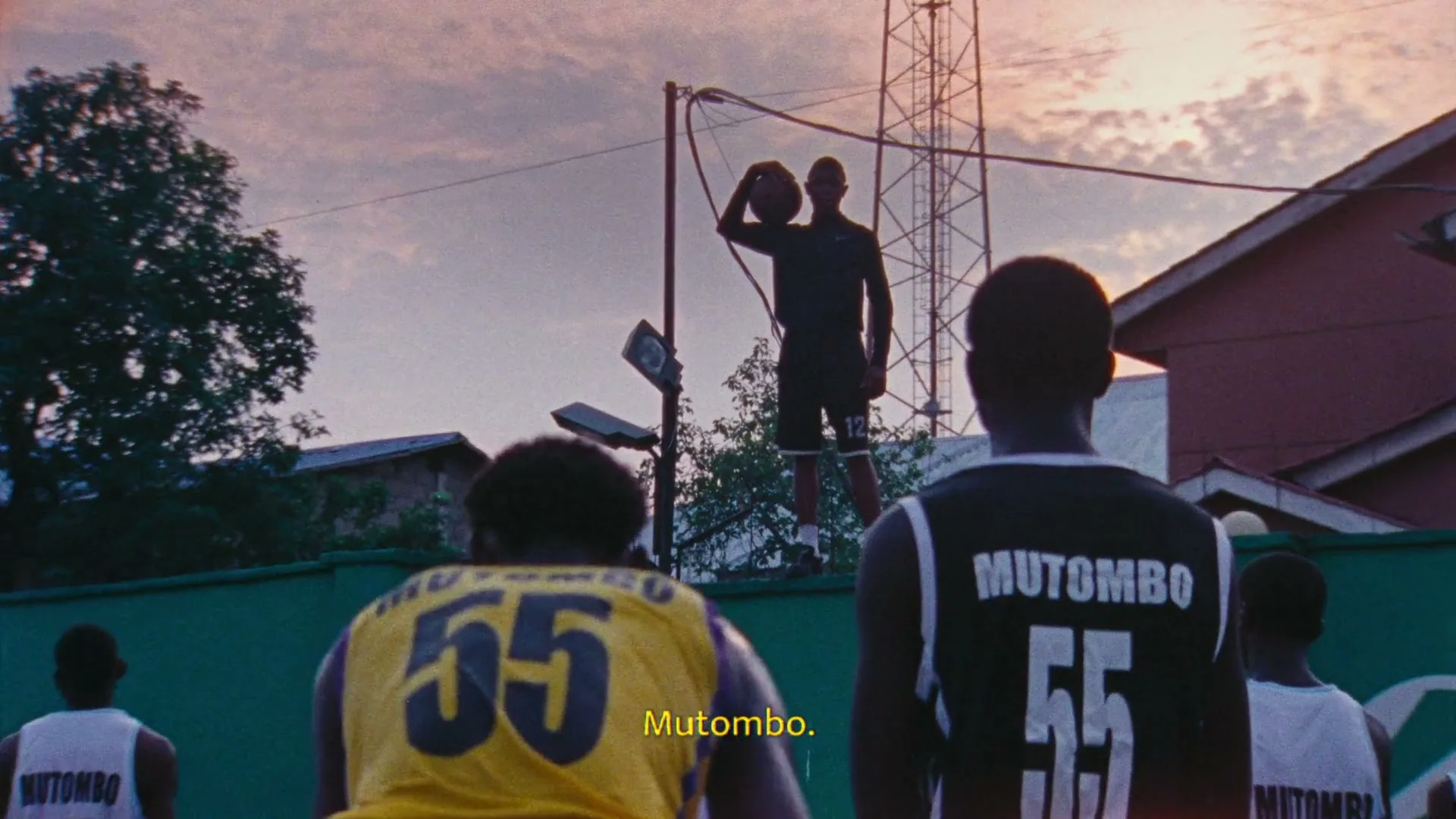 A young male basketball player in a black tracksuit holds a basketball on his shoulder, silhouetted against a sunset sky, as other players watch him.