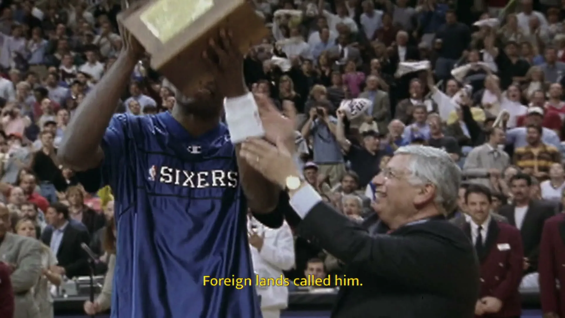 A male basketball player in a blue jersey receives a trophy from an older man in a suit, celebrating in a crowded arena.