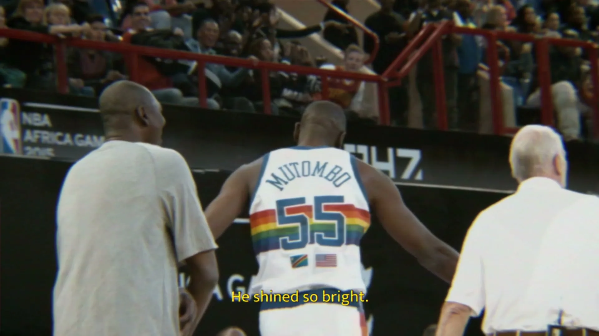 Dikembe Mutombo, a tall Black man in a white basketball jersey with "MUTOMBO 55" and a rainbow design, walks away from the camera on a basketball court, surrounded by other men and spectators.