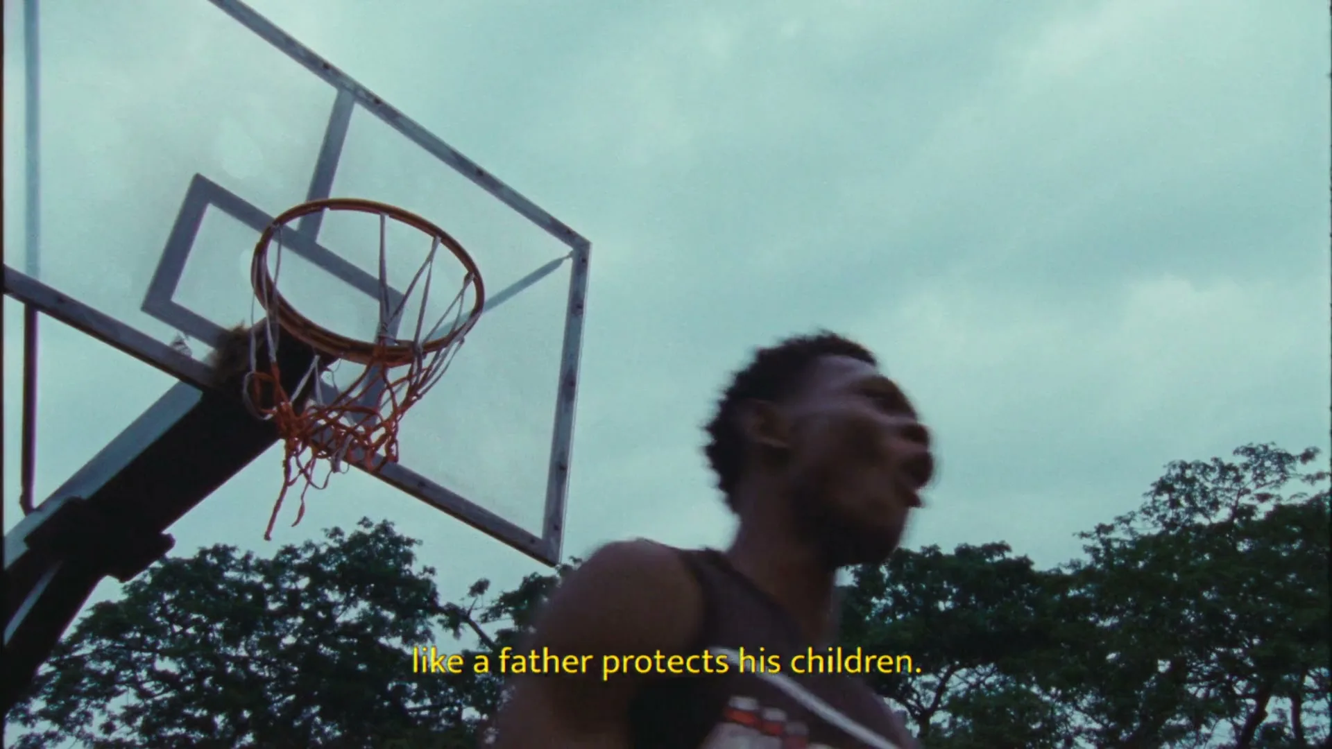 Two young men play basketball outdoors, one jumping to shoot a ball towards the hoop, while the other defends.