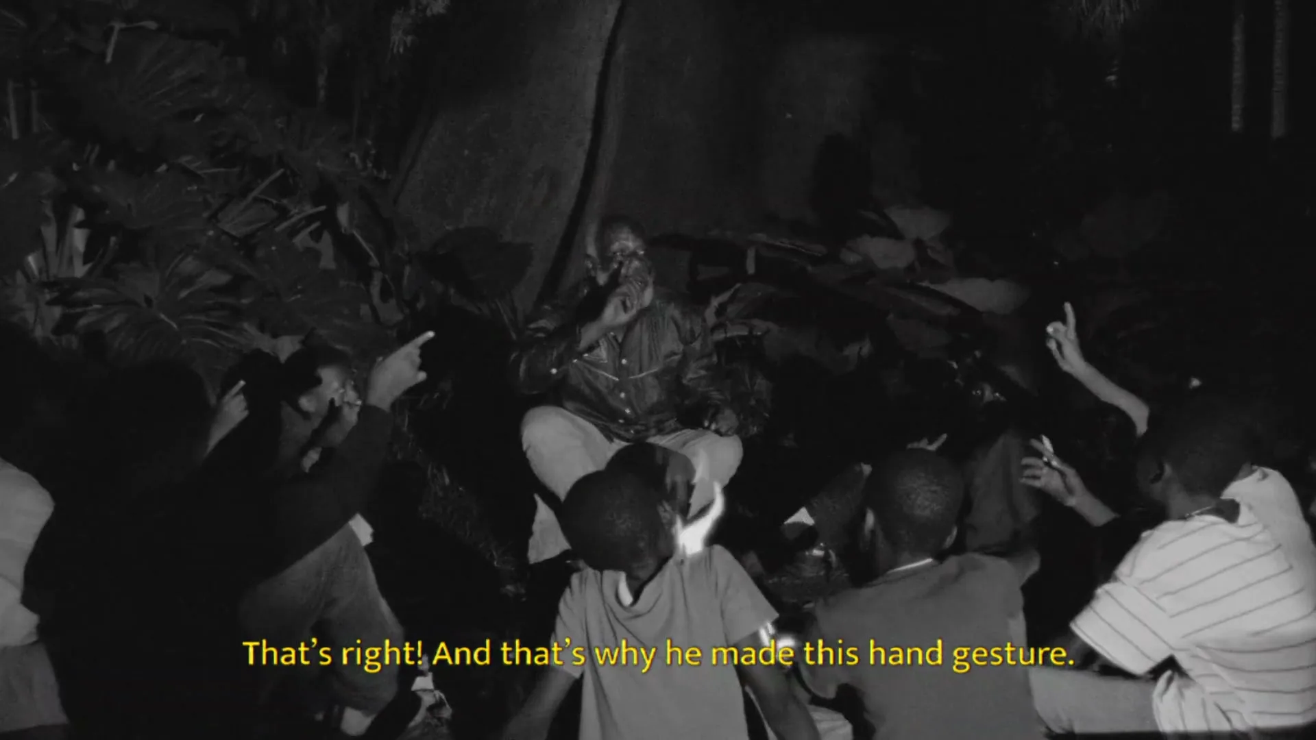 A group of young men sit around an older man who is speaking and gesturing with his hands in a dark, outdoor setting at night.
