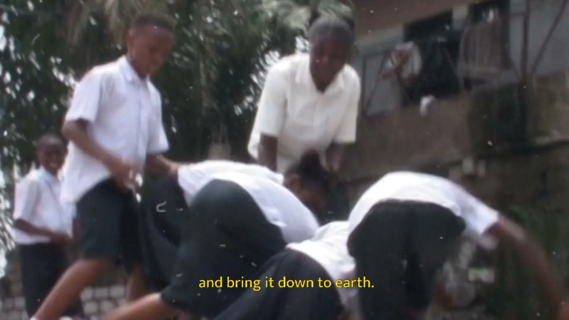 A group of children in school uniforms play a rough game, possibly rugby or wrestling, on a dirt mound outdoors during the day.