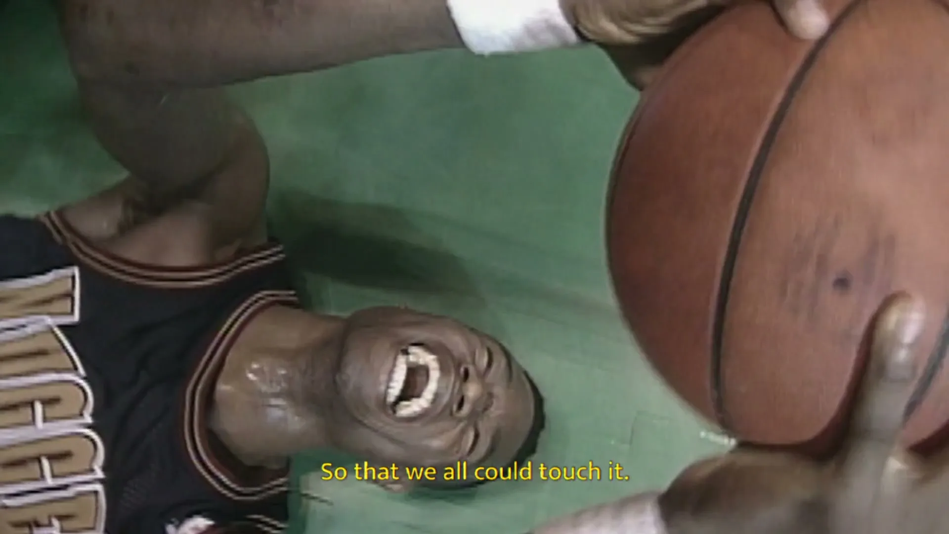 A basketball player in a blue jersey lies on a green court, screaming in agony, while another player's hand holds a basketball above him.