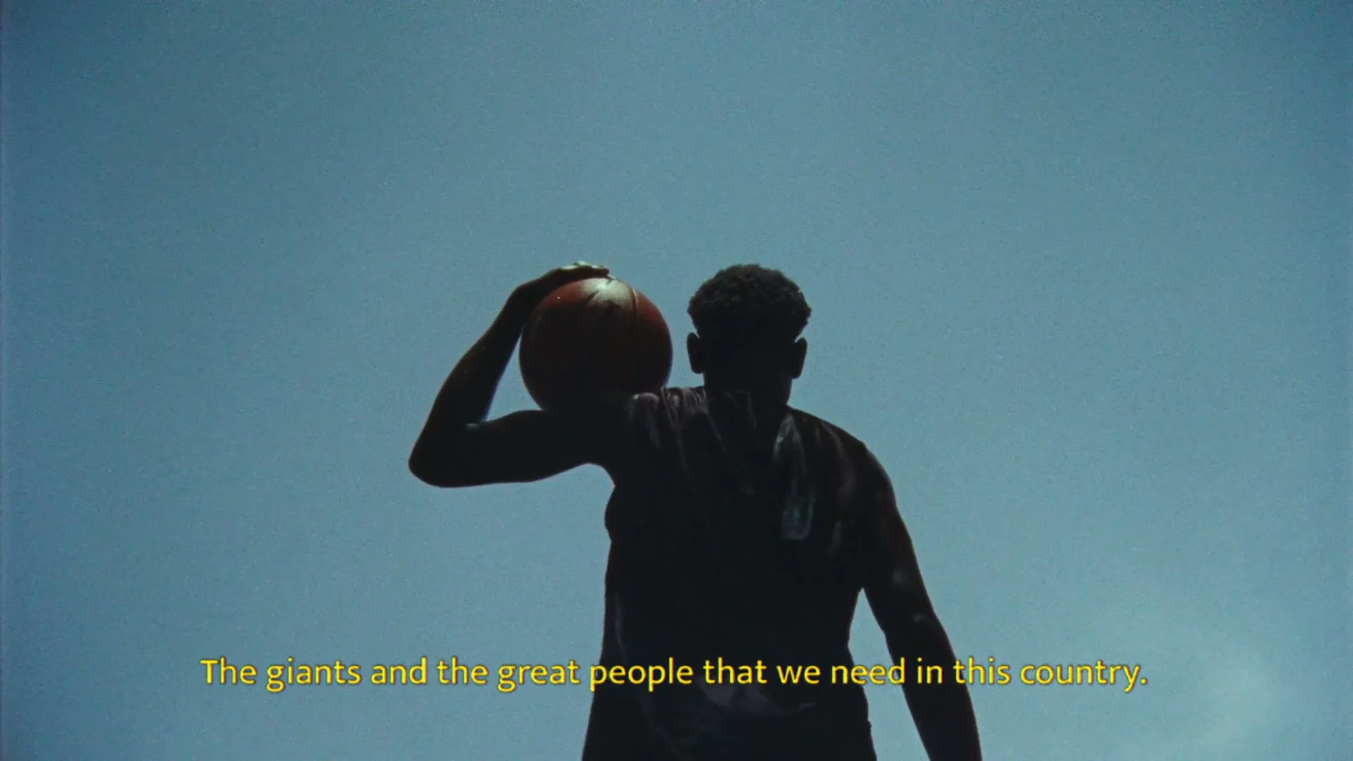 A male basketball player, silhouetted against a bright sky, holds a basketball over his head, with yellow text at the bottom of the frame.