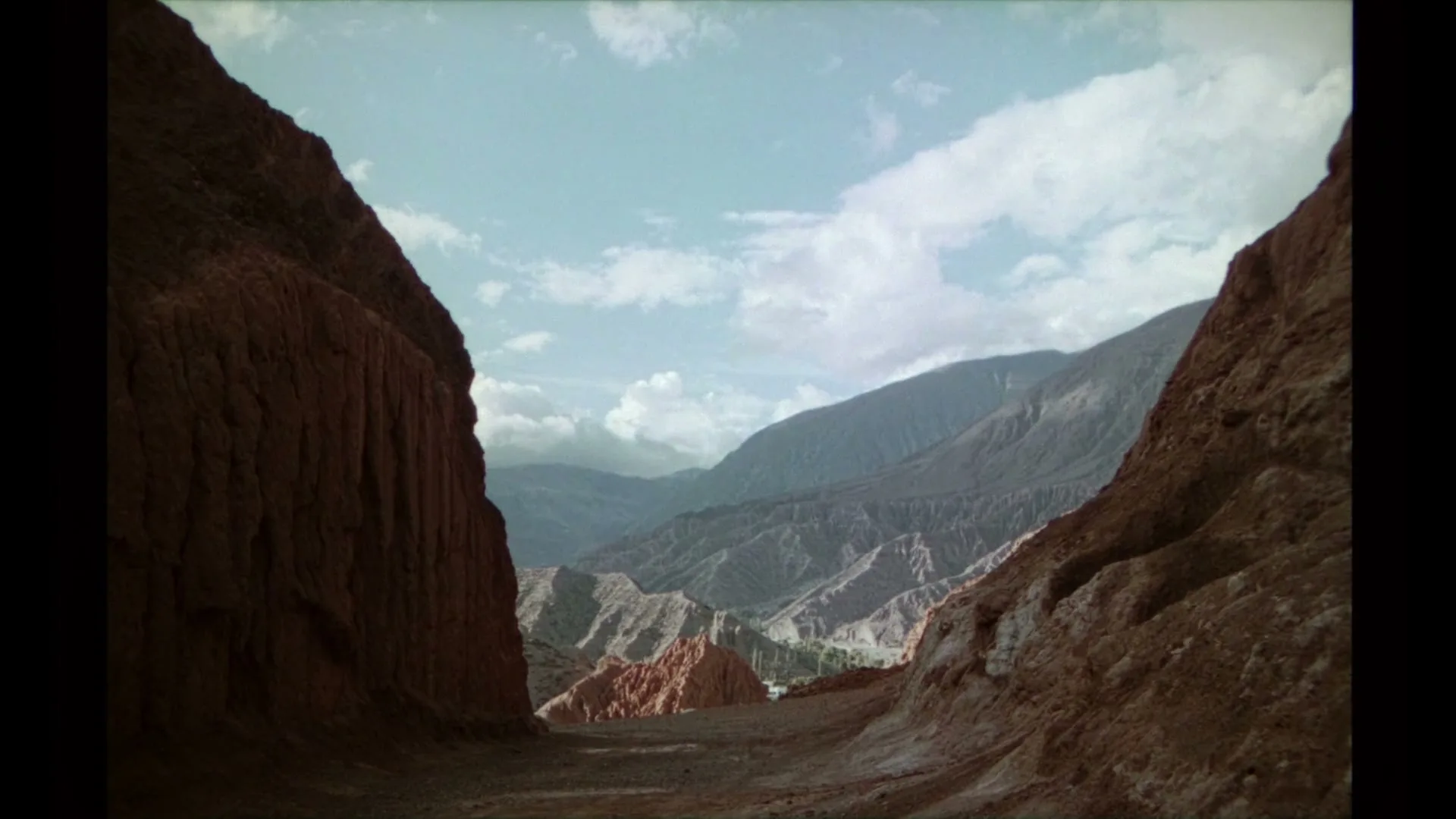 A wide shot of a vast, rugged canyon with towering red rock formations on either side, leading to distant mountains under a partly cloudy sky.